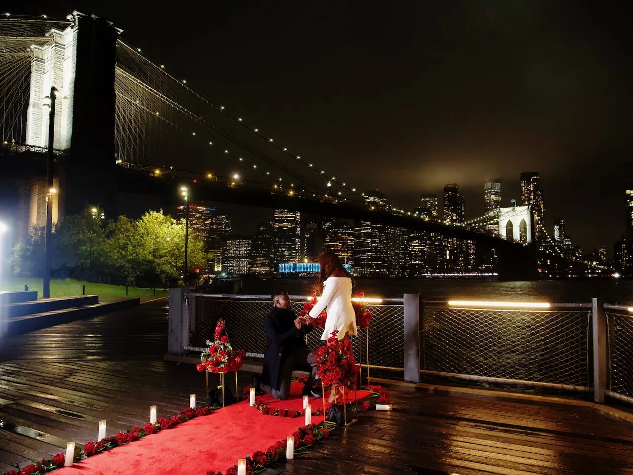 Proposal shoot: a man proposes to a woman on a red carpeted platform decorated with flowers and candles, with the Brooklyn Bridge and New York City skyline illuminated in the background at night.