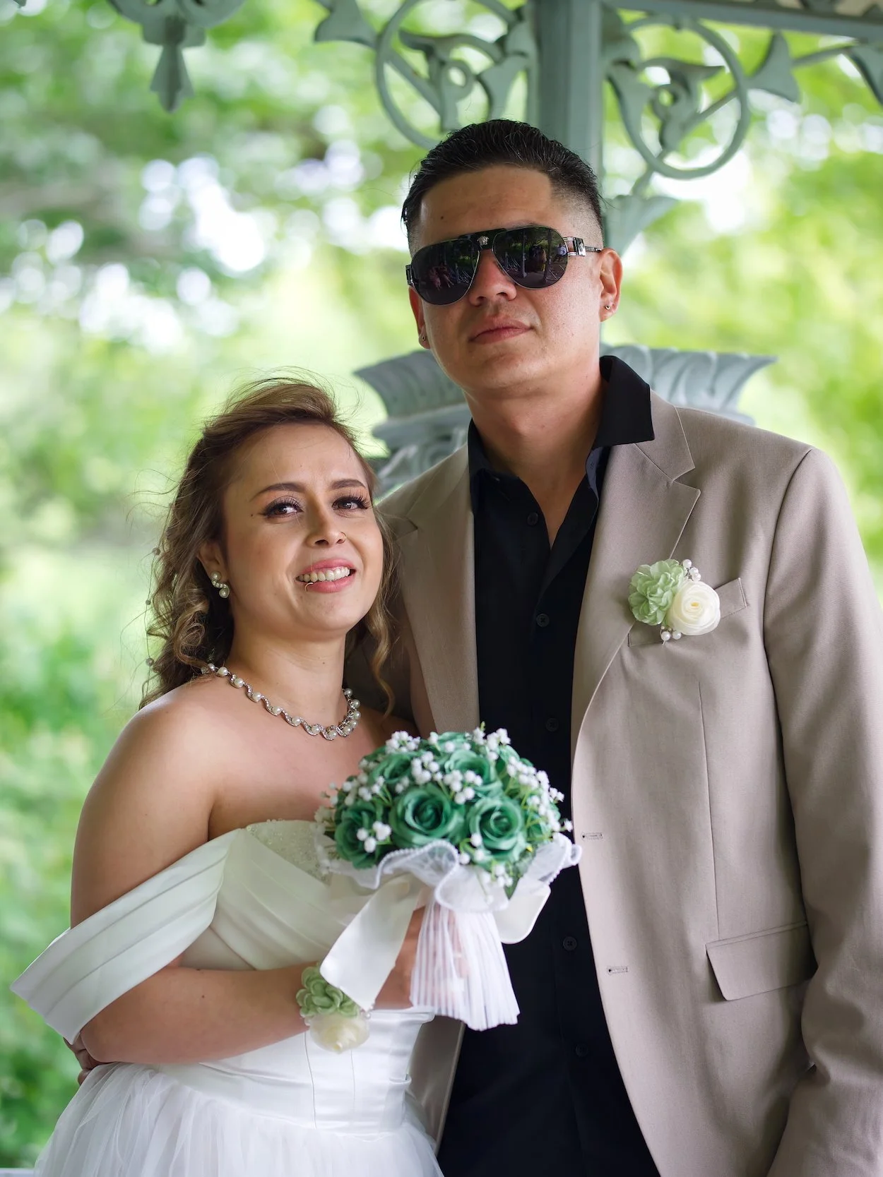 Wedding Portrait, Central Park: A bride and groom posing outdoors, the bride in a white wedding dress holding a bouquet, and the groom in a beige suit with black shirt and sunglasses, under a green leafy canopy.