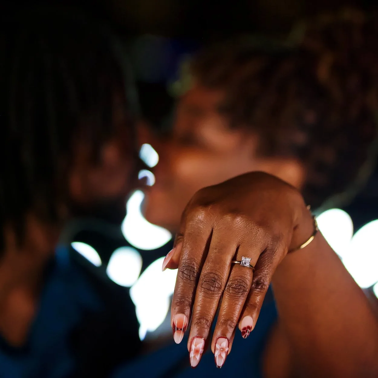 Proposal shoot: close-up of a woman's hand with a diamond engagement ring, showing intricate red and white nail art, in front of a blurred background of a woman with curly hair.