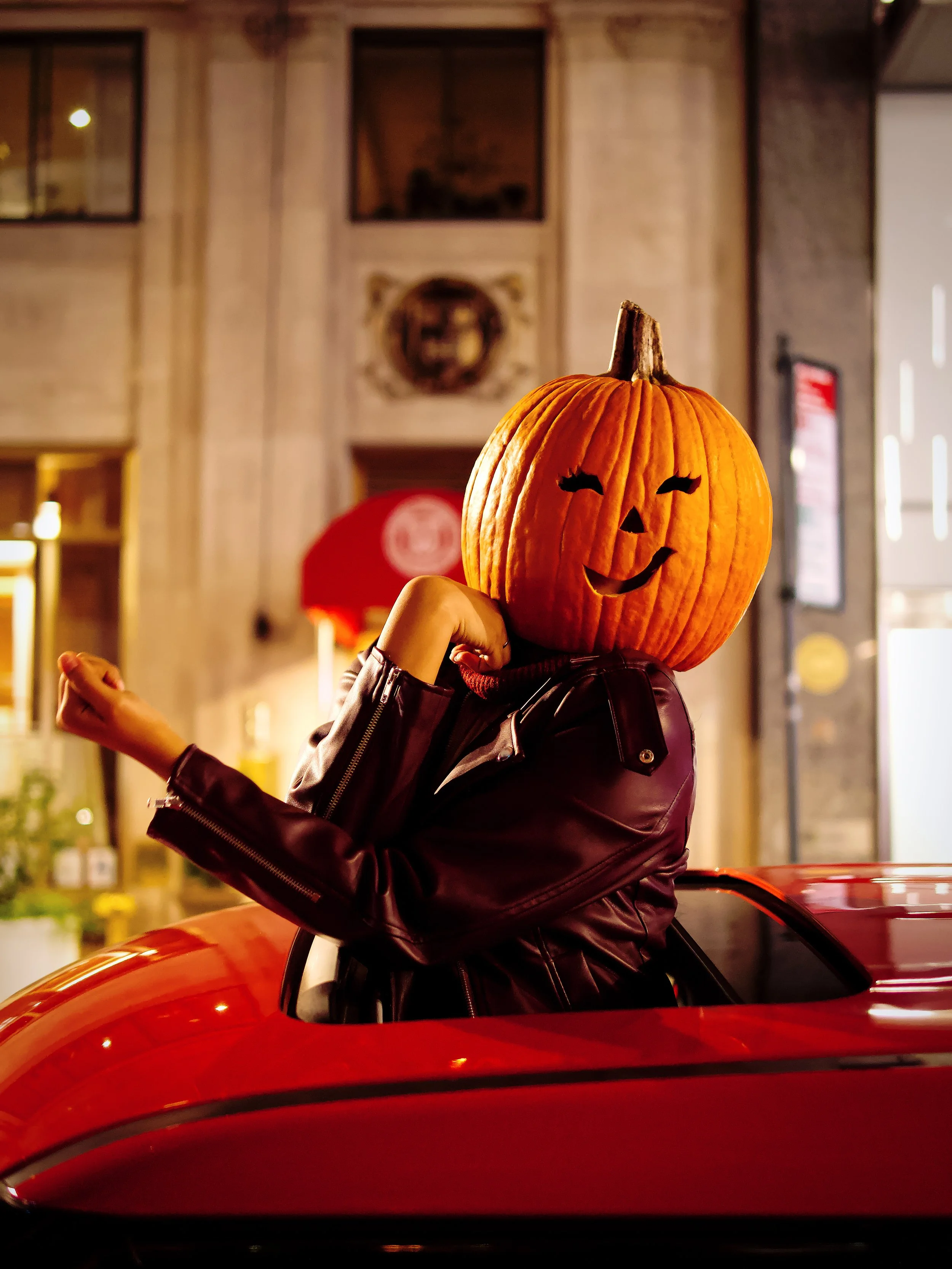 Creative fall and Halloween lifestyle portrait of a person wearing a carved pumpkin head and leather jacket in a car sunroof during a fun NYC seasonal photoshoot.
