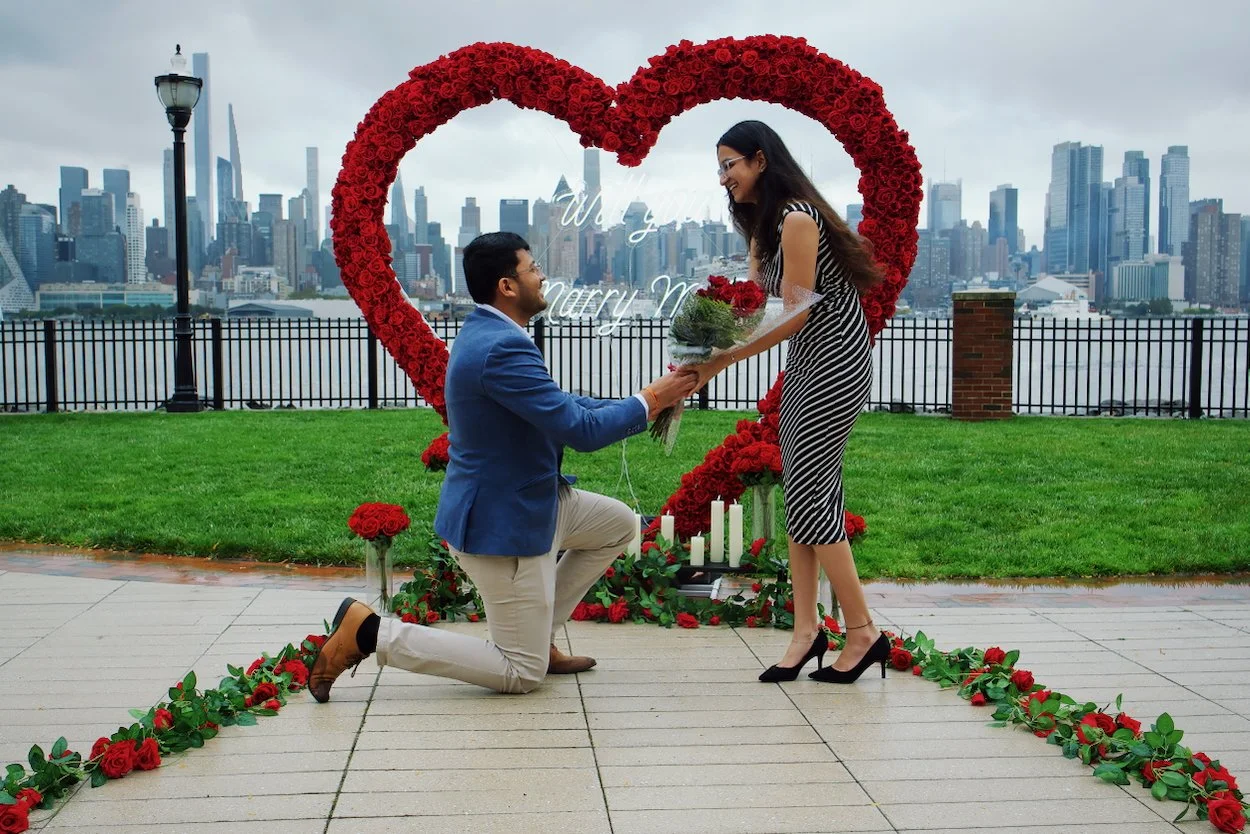 A man proposing marriage to a woman in front of a heart-shaped floral display with a city skyline in the background. She is holding a bouquet of red roses, smiling, and dressed in a striped dress. The man is kneeling on one knee, holding her hands, d