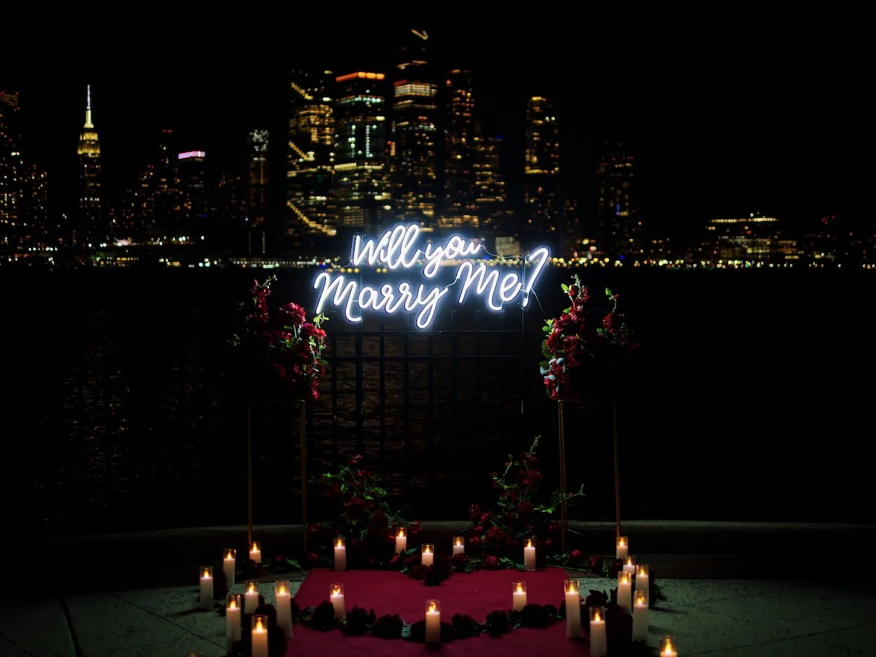 A nighttime cityscape with tall buildings illuminated by lights, and a neon sign that says "Will you marry me?" surrounded by flowers and candles on the ground.