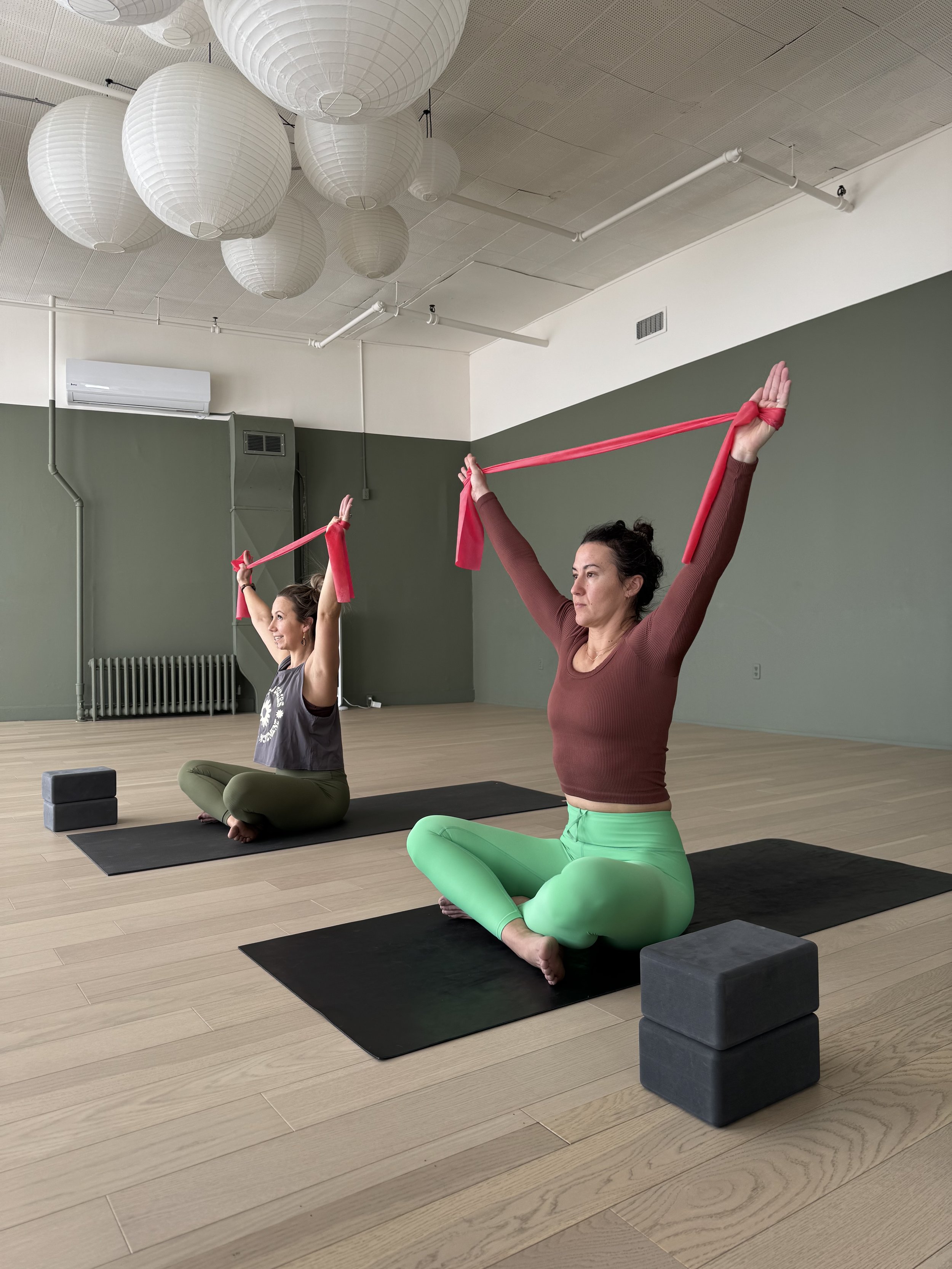 Two women practicing yoga indoors, sitting cross-legged on black mats, holding pink resistance bands overhead, with yoga blocks nearby