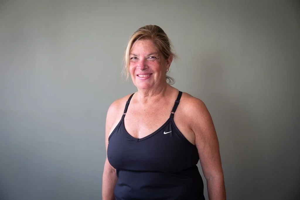 A middle-aged woman with blonde hair tied back, wearing a black Nike tank top, standing against a plain light gray wall, smiling at the camera.