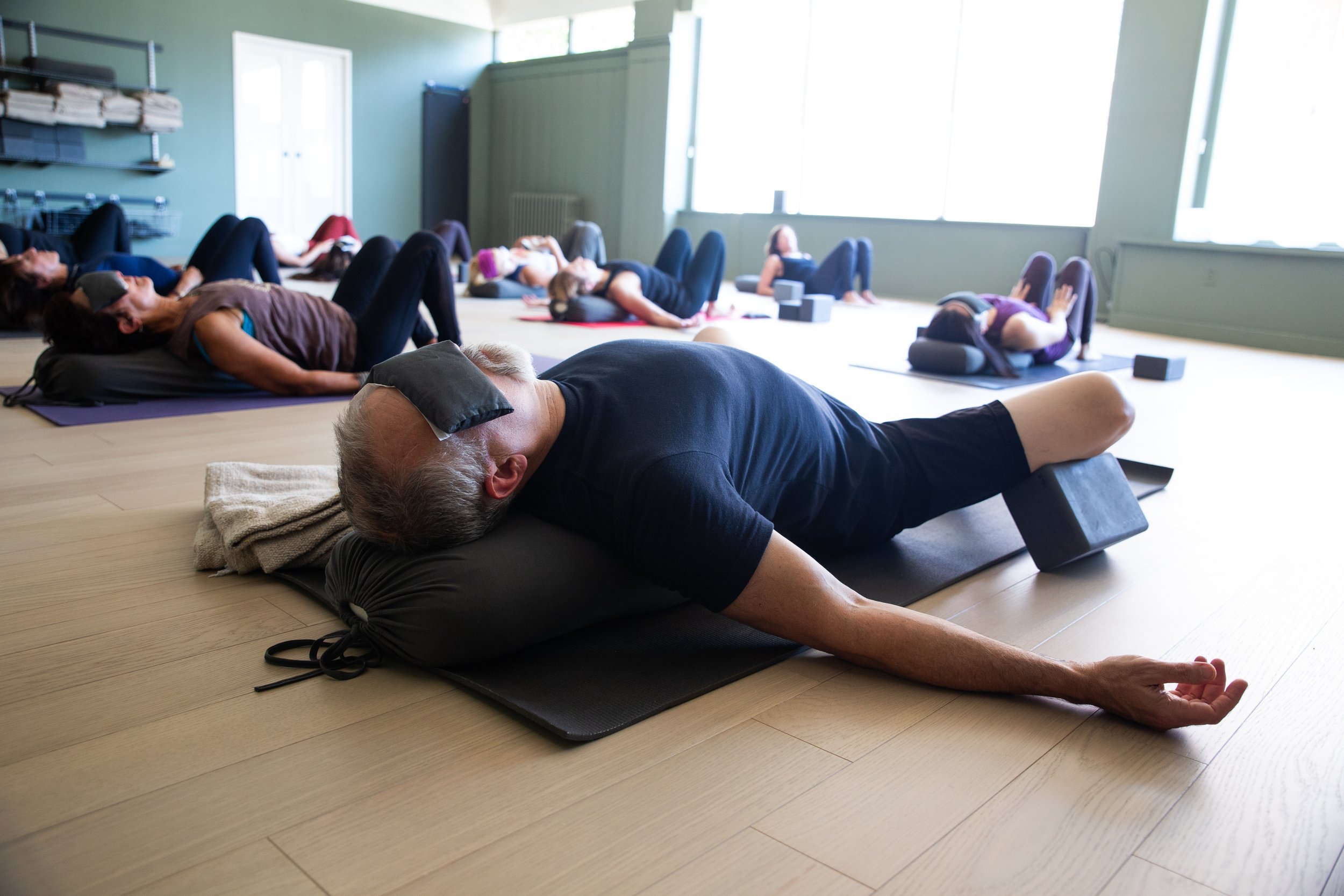A group of people in a yoga class are lying on mats, practicing relaxation and stretching exercises in a bright room with large windows.