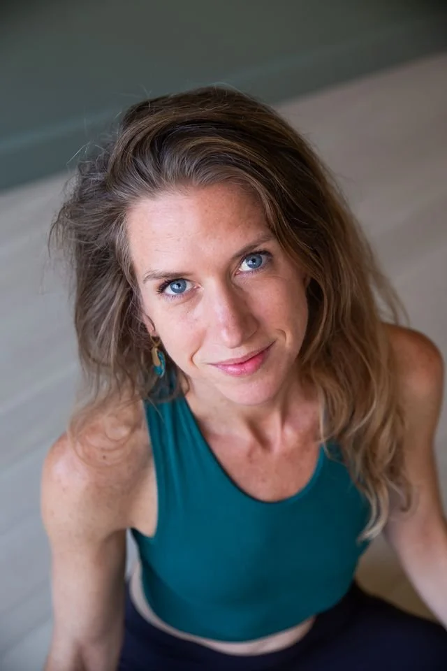 A woman with long wavy hair and blue eyes, wearing a teal sleeveless top, smiling and looking up at the camera.