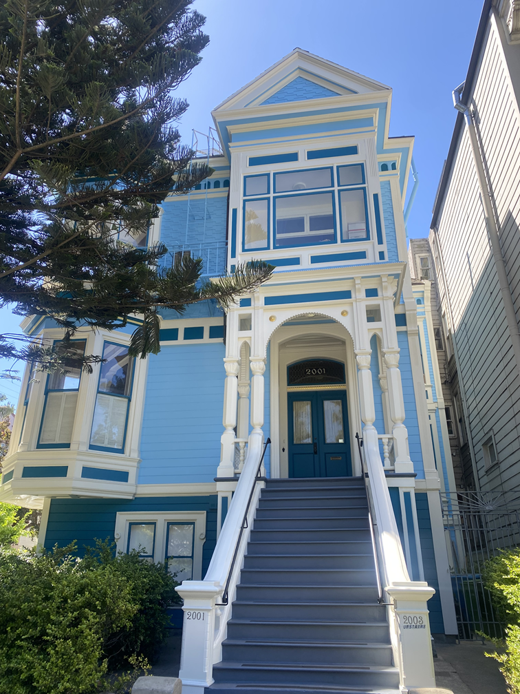Blue Victorian-style house with white trim and stairs leading to the front door, surrounded by greenery and a large tree.