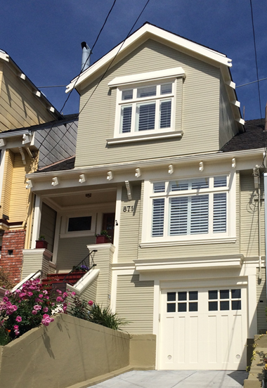A multi-story house with a light-colored exterior, bay windows, and a garage door, with colorful flowers and a small staircase leading to the front porch.