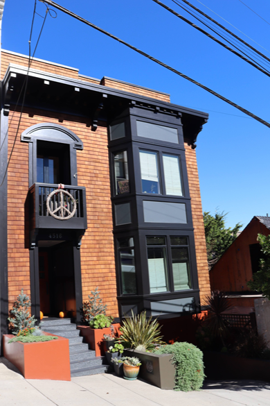 Exterior of a modern brick building with a black bay window and a small balcony with a peace sign decoration, surrounded by potted plants and a staircase leading to the entrance.