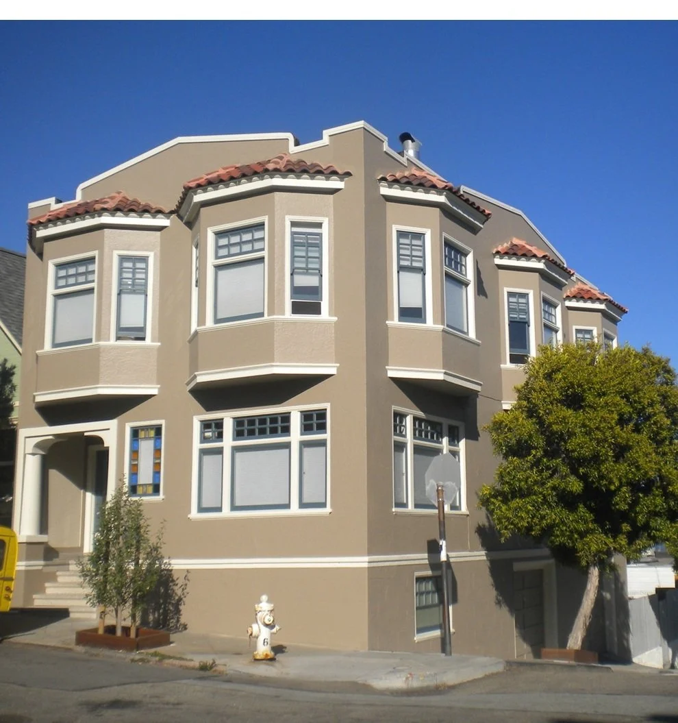 A three-story beige residential building with red-tiled roof, bay windows, a small front yard with a tree, and a fire hydrant on the sidewalk during a sunny day.