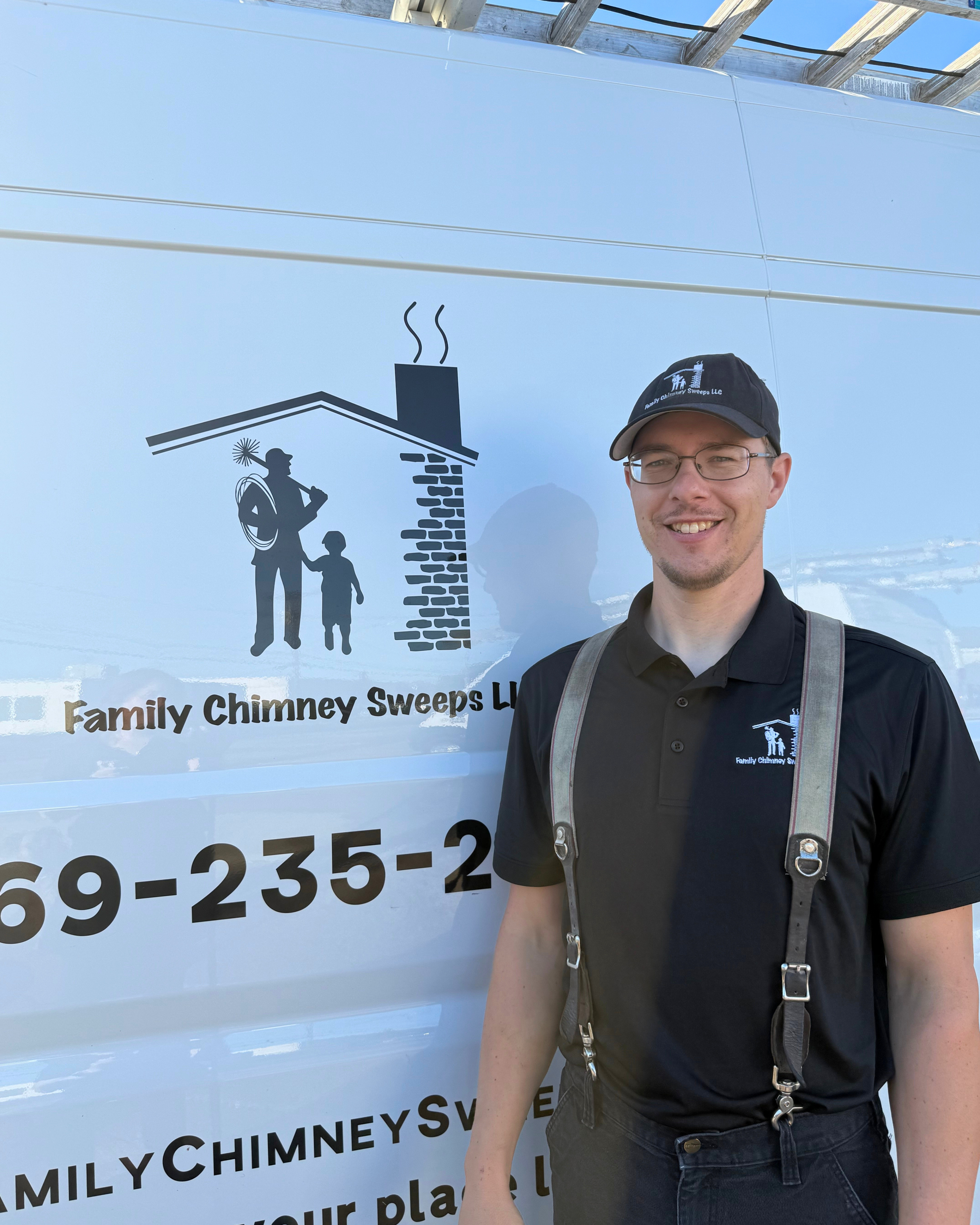 Young man with glasses smiling, wearing a black polo shirt with a company logo, standing in front of a white van with a family chimney sweep logo and contact information, holding a chimney sweep tool over his shoulder.