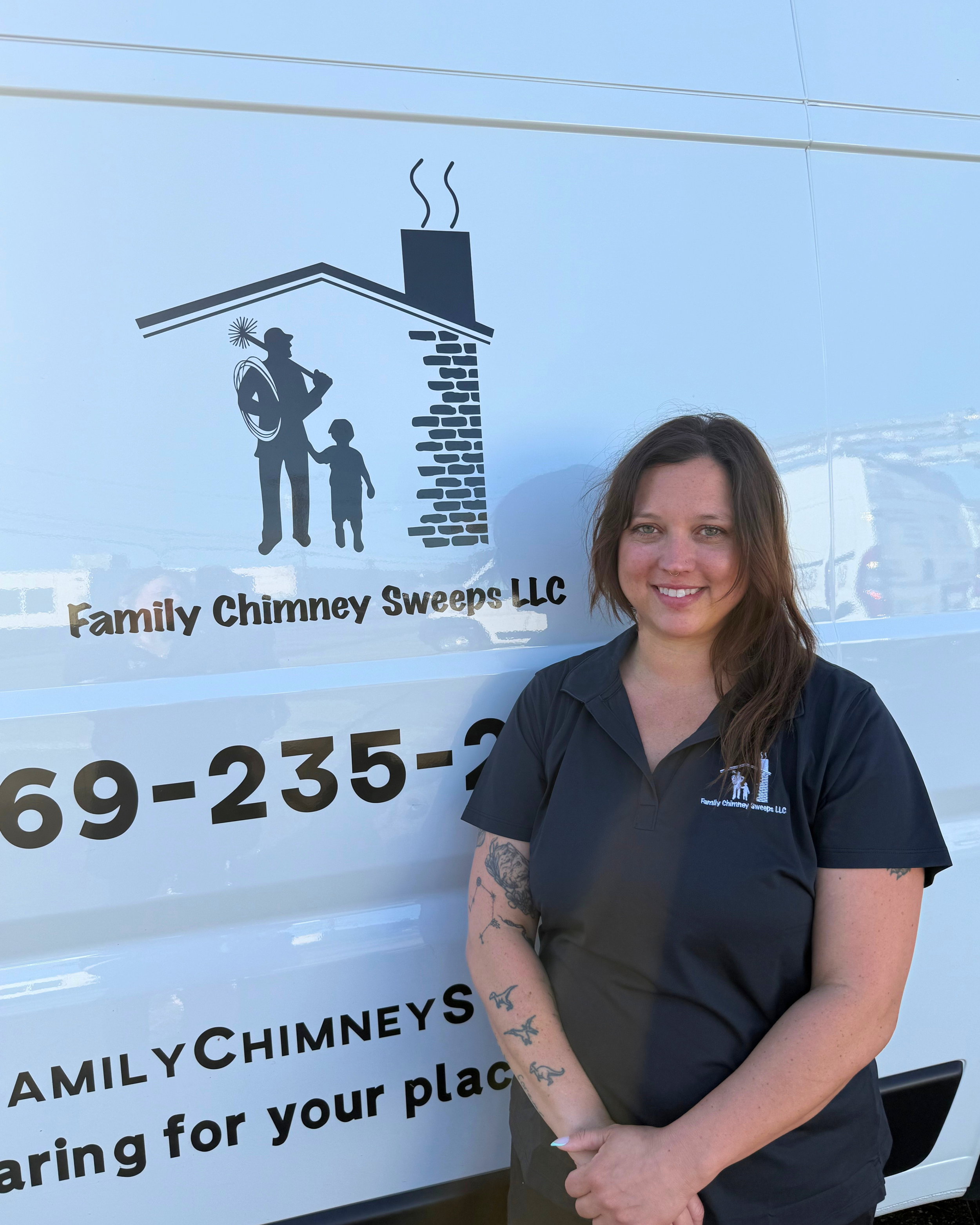 A woman with glasses and long hair smiling in front of a white van with a chimney sweep company logo. The van has the company name 'Family Chimney Sweeps LLC' and a phone number. She is wearing a navy shirt with the same logo.