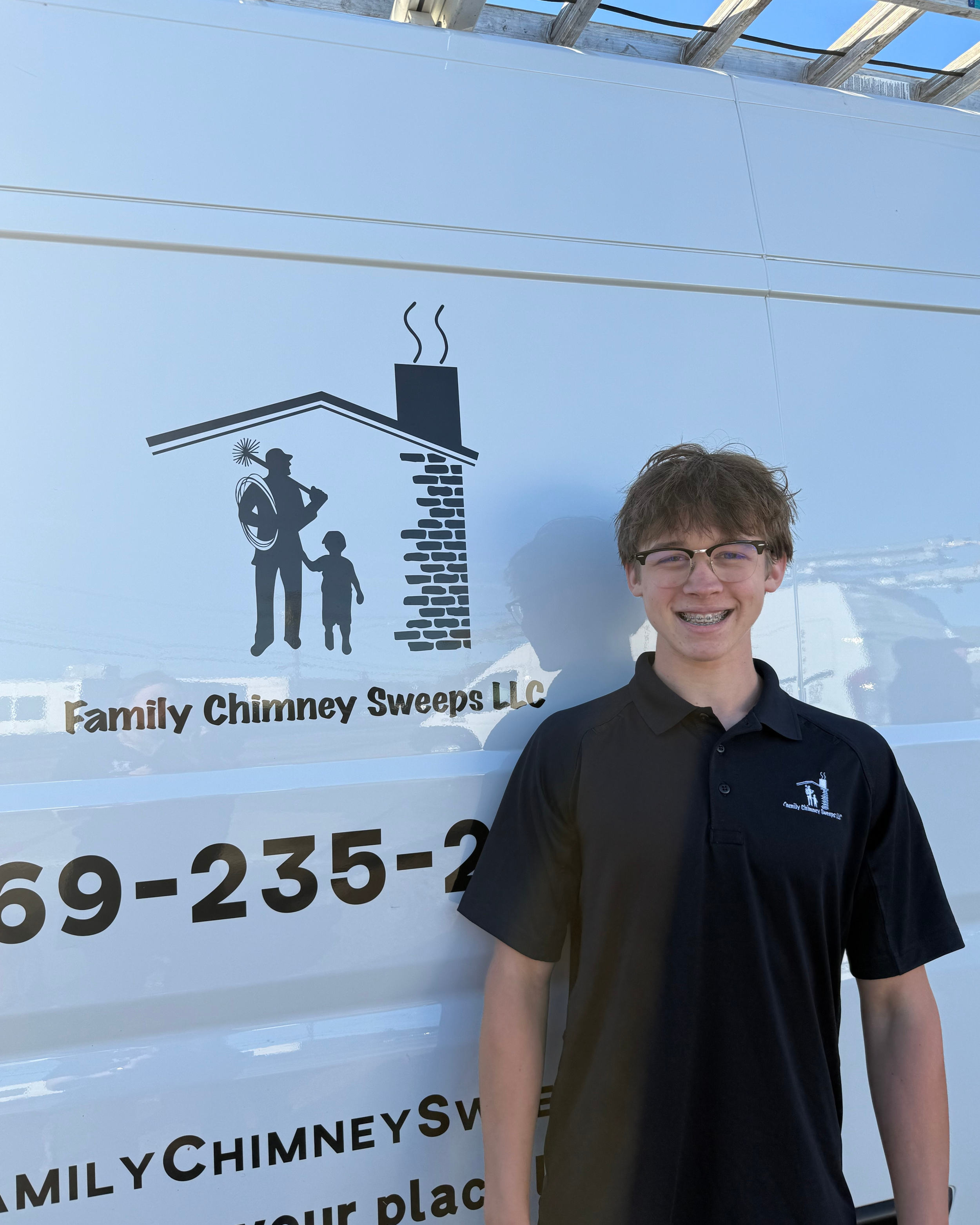 Young man standing outdoors in front of a chimney sweep company's truck, wearing a navy polo shirt with the company logo.