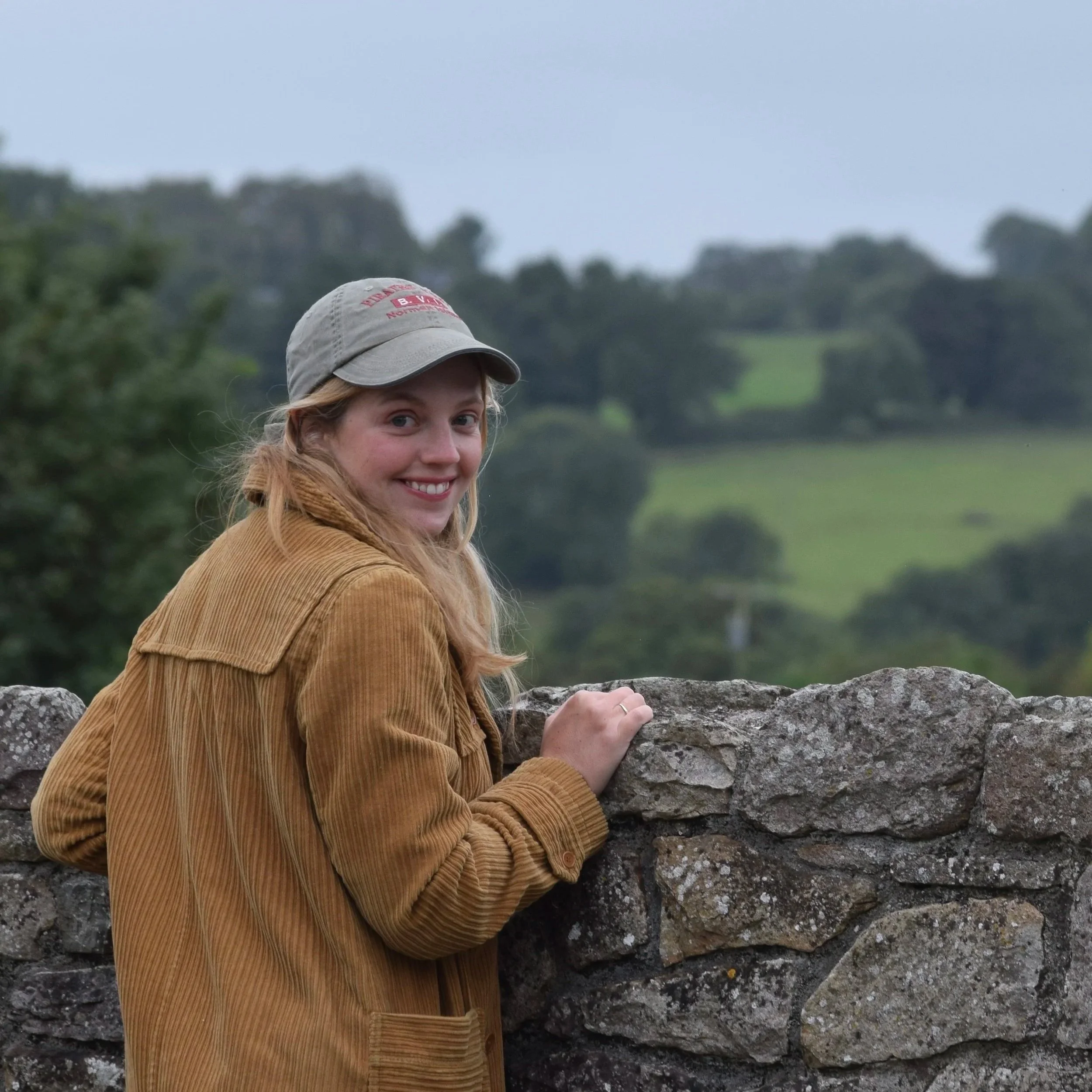 A woman with red hair and a beige hat smiling while standing outdoors next to a stone wall, with a background of green trees and a cloudy sky.