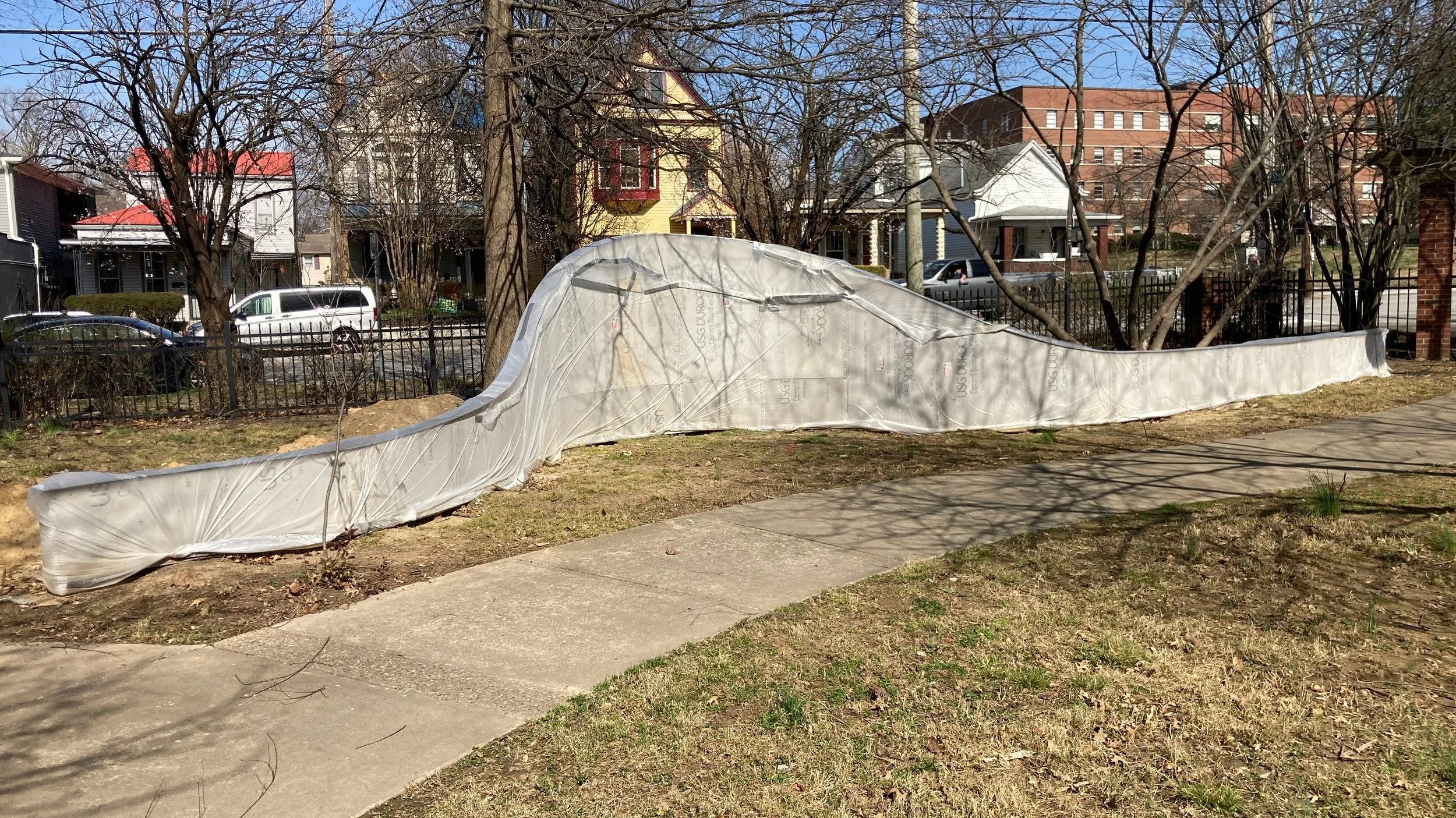 Just behind a curving concrete walkway in a park, a serpentine "wall" structure stands, covered in protective plastic. The structure is about 30 feet long, lowest at either end, and highest in the middle. It curves in a gentle arc.