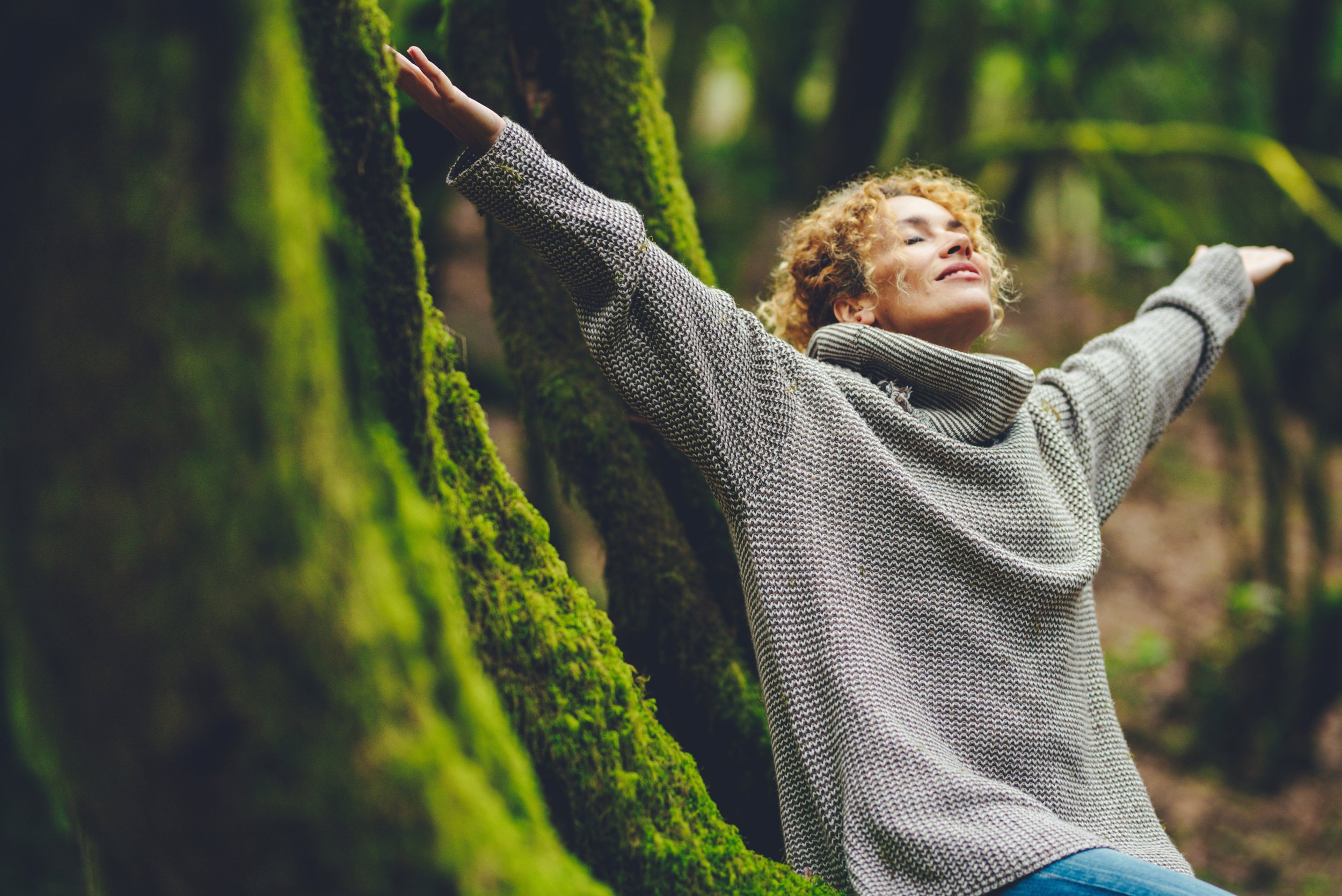 Light brown curly haired smiling woman with outstretched arms embracing the sun in a green mossy tree grove. closed eyes, serene smile