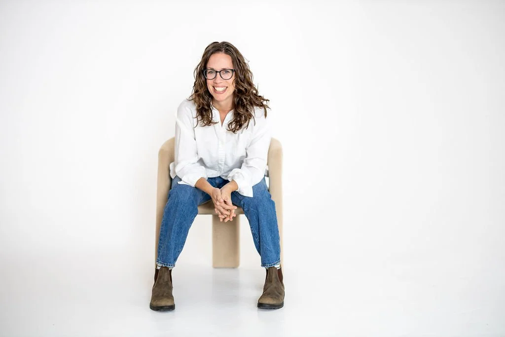 Image of woman sitting on chair with hands-on her knees. She is wearing a white button up shirt, blue jeans and brown boots.