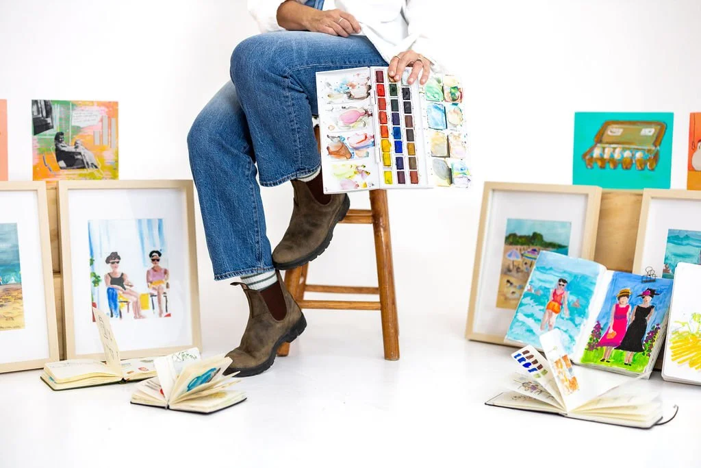 Photo of an artist sitting on a stool holding a watercolor palette and paintings and journal behind the stool.