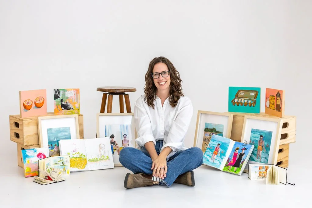 Women with brown curly hair sitting criss cross in front of a few of her colorful paintings and journals placed behind her.