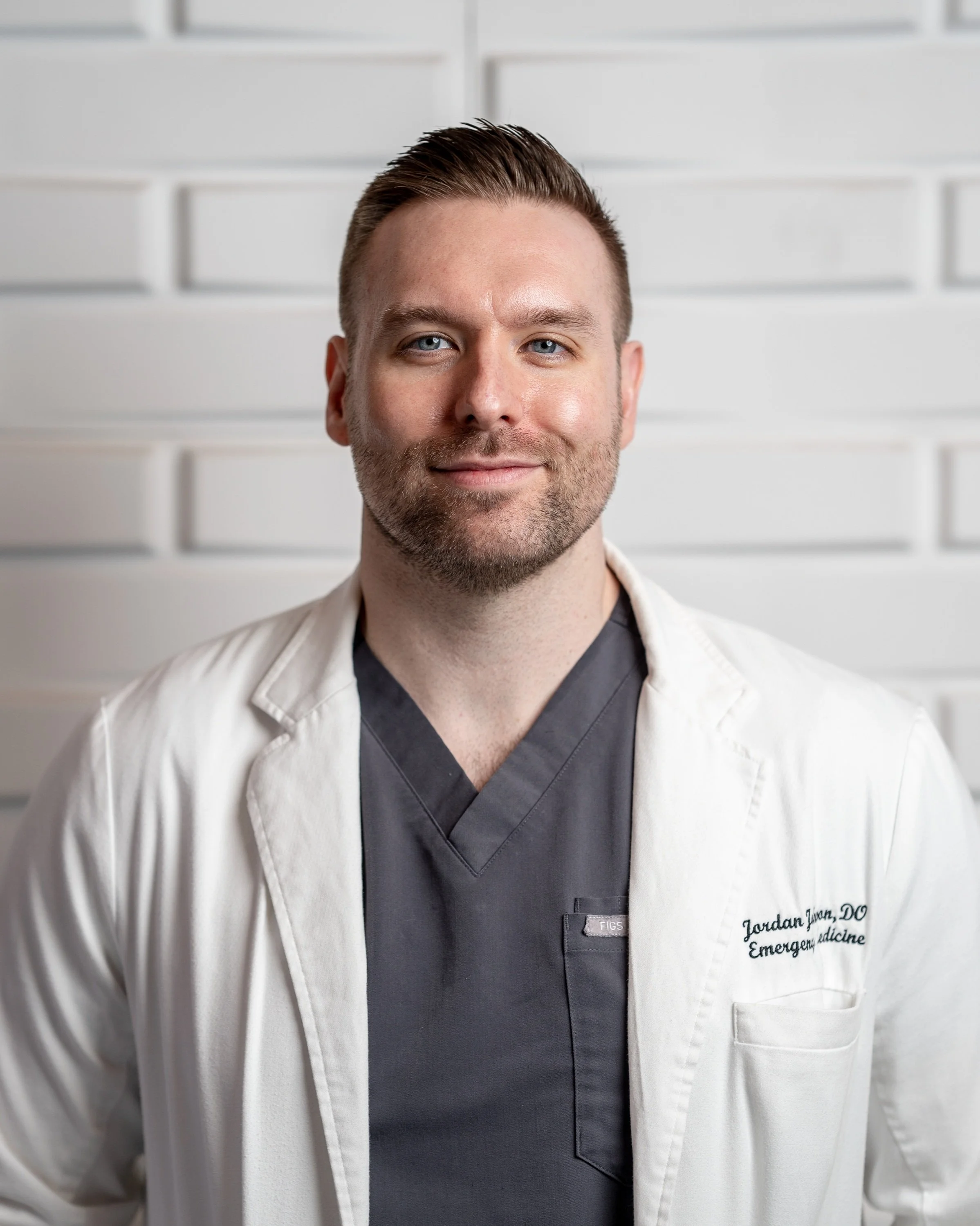 A male doctor wearing a white coat and grey scrubs, standing against a white wall with geometric patterns.