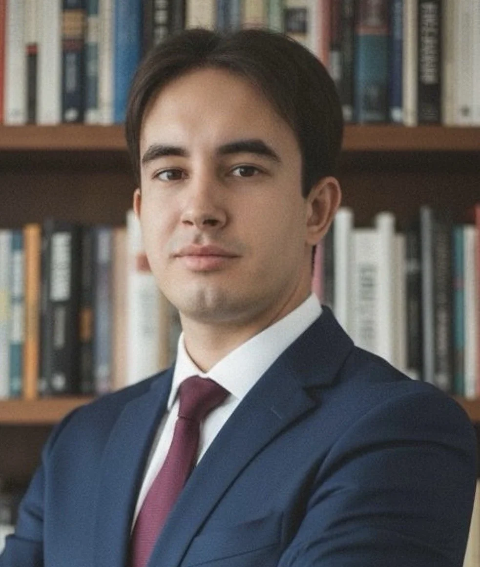 Portrait of a young man with dark hair, wearing a grey suit and black shirt, against a plain grey background.