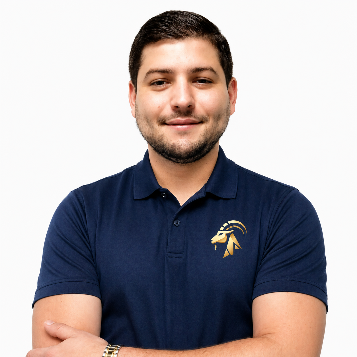 A young man with short dark hair and a beard, smiling, wearing a light blue polo shirt with a logo on the chest, standing in front of a plain, light-colored wall.