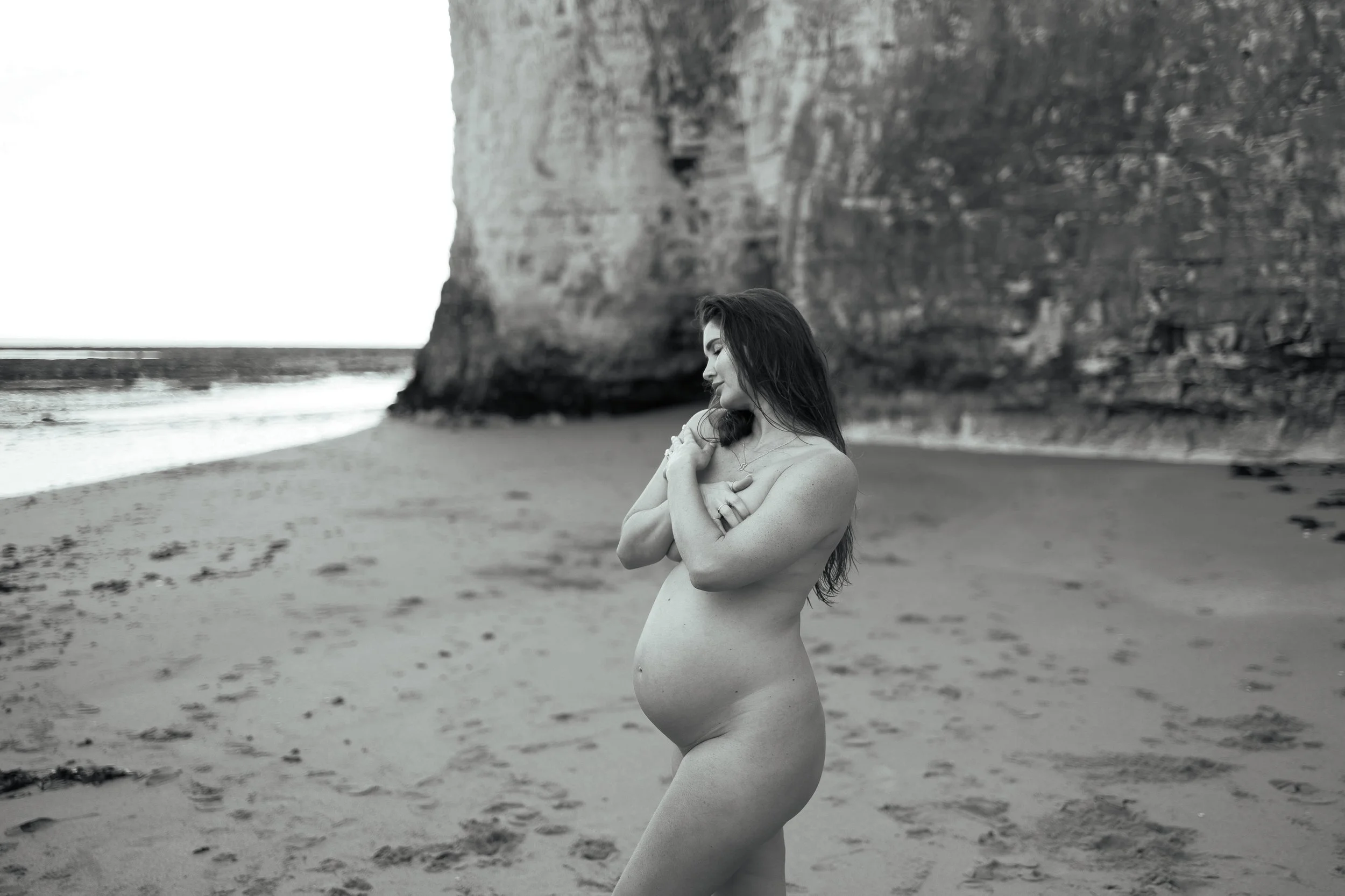 A pregnant woman standing on a beach near a large cliff, embracing herself with her arms crossed over her chest, looking down with a peaceful expression, in black and white.