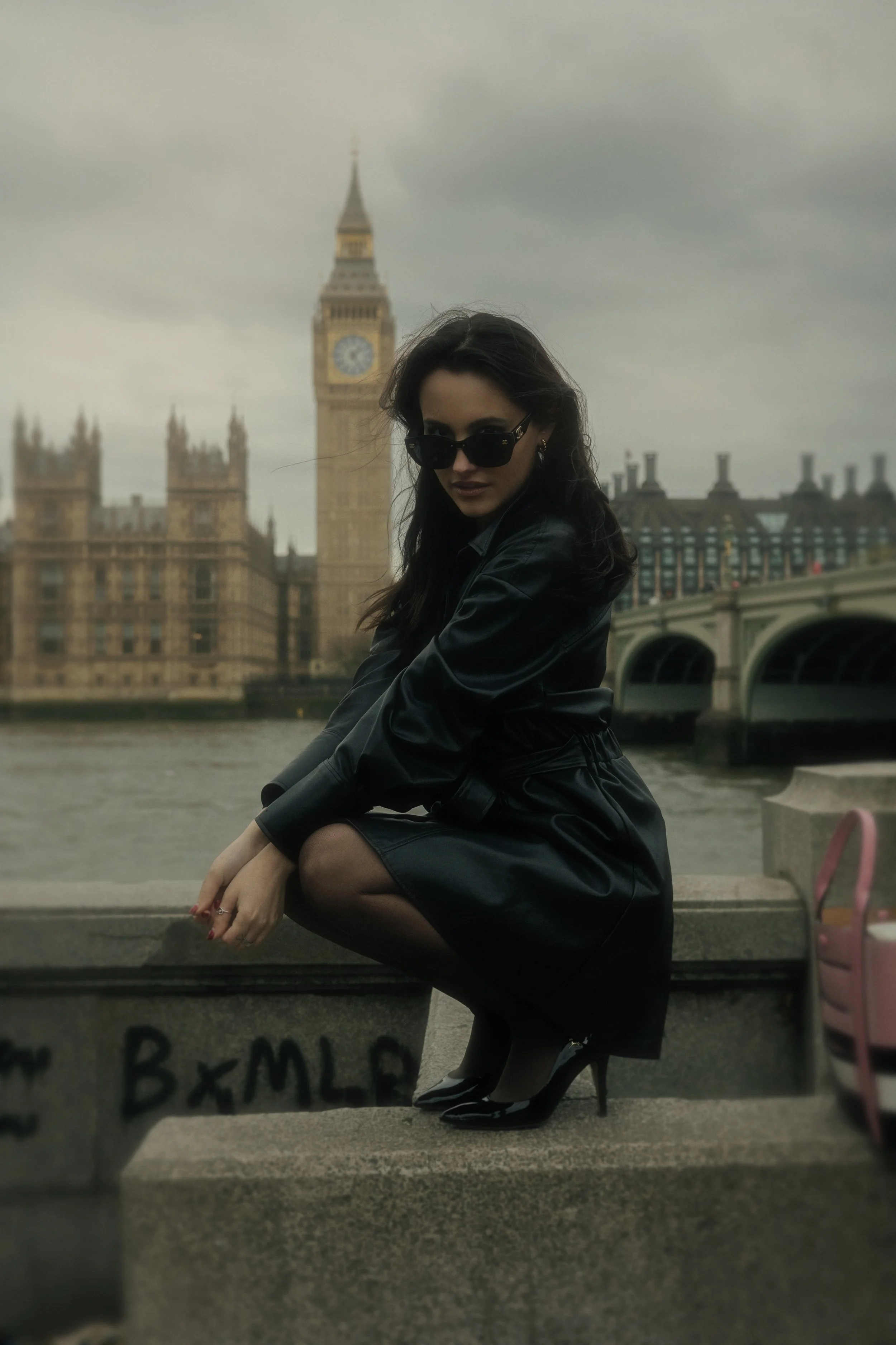 A woman wearing sunglasses, a black leather jacket, and black high heels squatting on a concrete ledge near the river with the Big Ben clock tower and the Palace of Westminster in London in the background, under a cloudy sky.