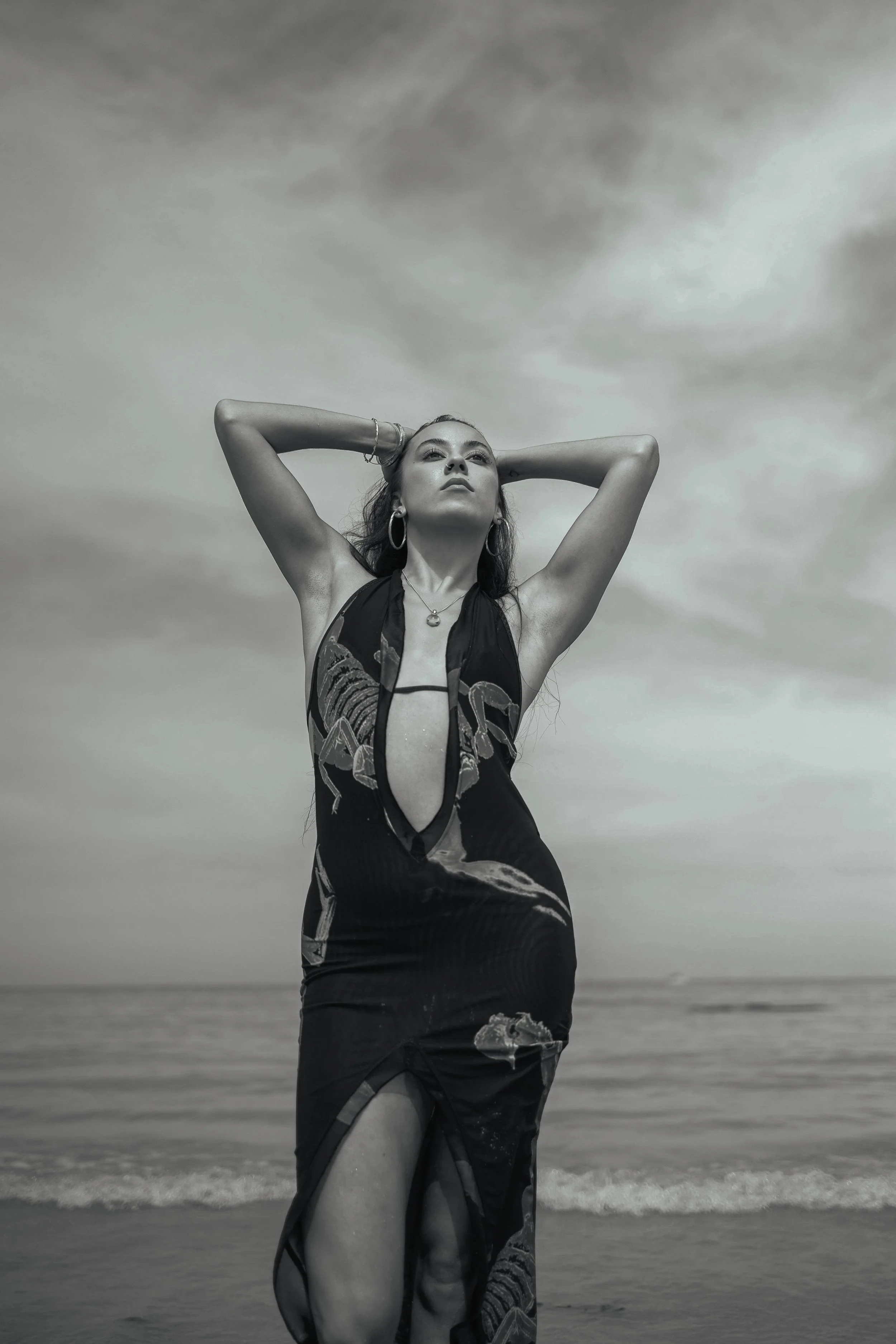 A woman standing on the beach with her hands behind her head, wearing a black dress with a snake print, and looking up. It's a black and white image with cloudy sky background.
