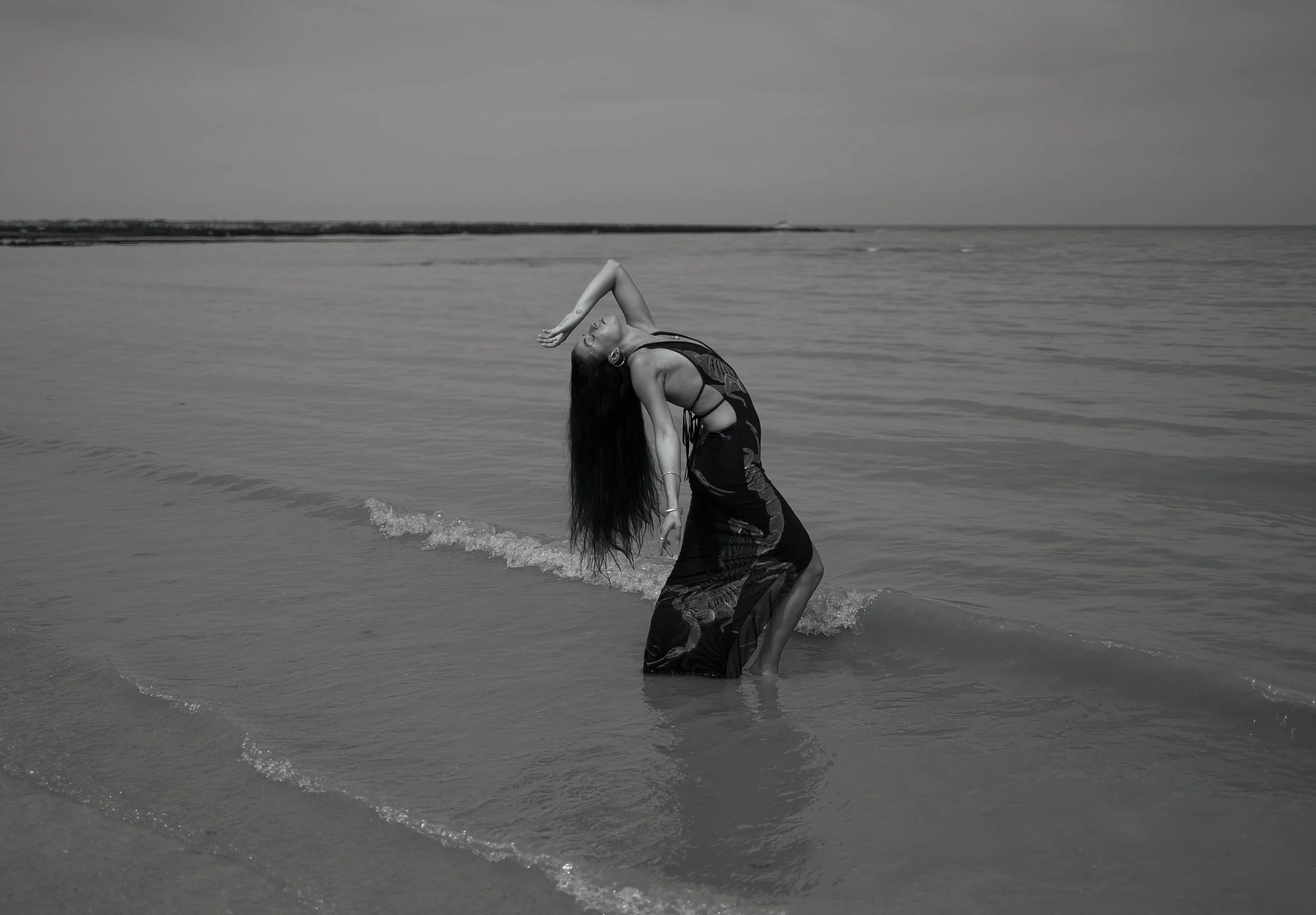 A woman in a long dress wading in the ocean with her head tilted back and arm raised, standing in shallow water near the shoreline.