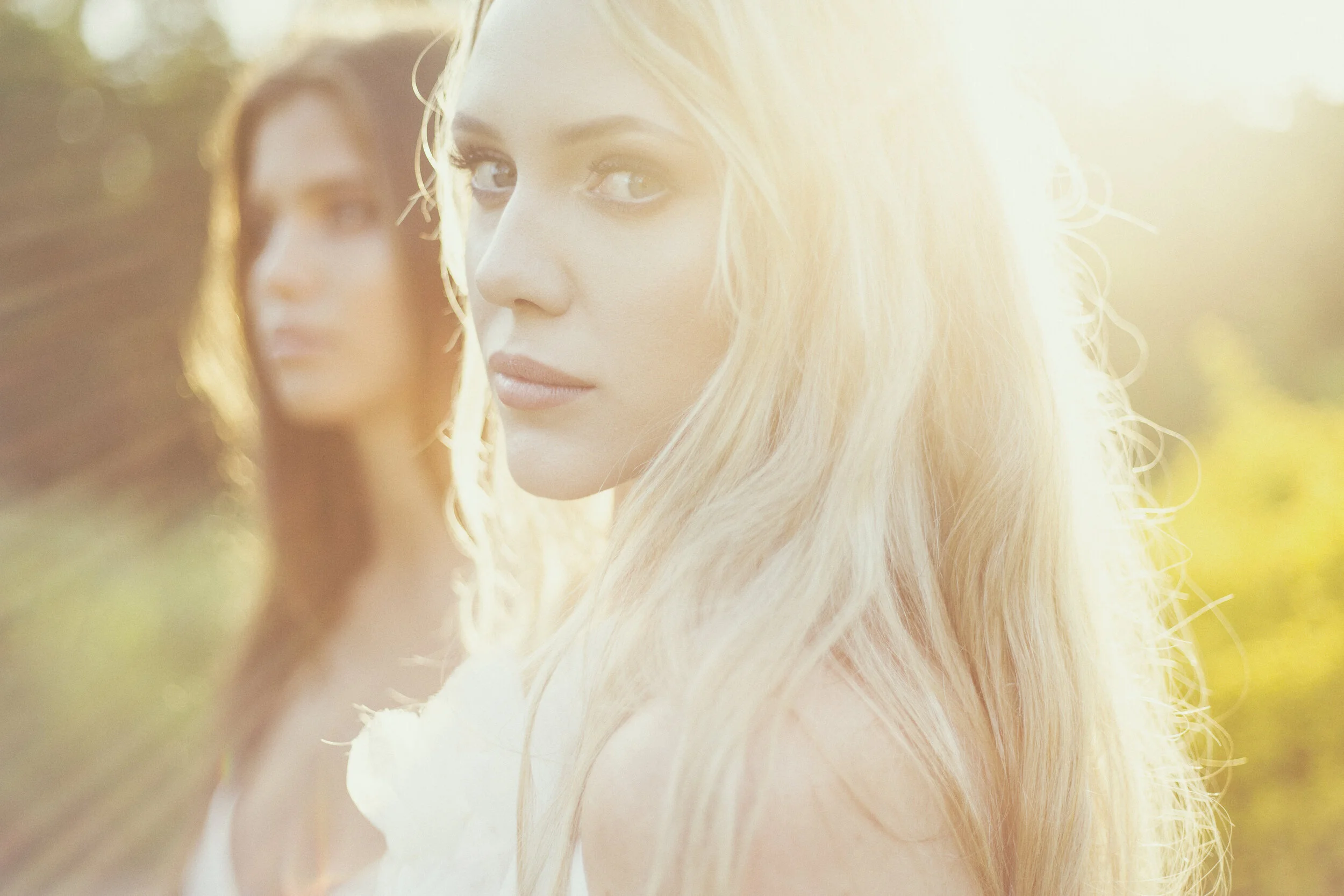 Two women with long hair standing outdoors in sunlight, with the woman on the right in focus.