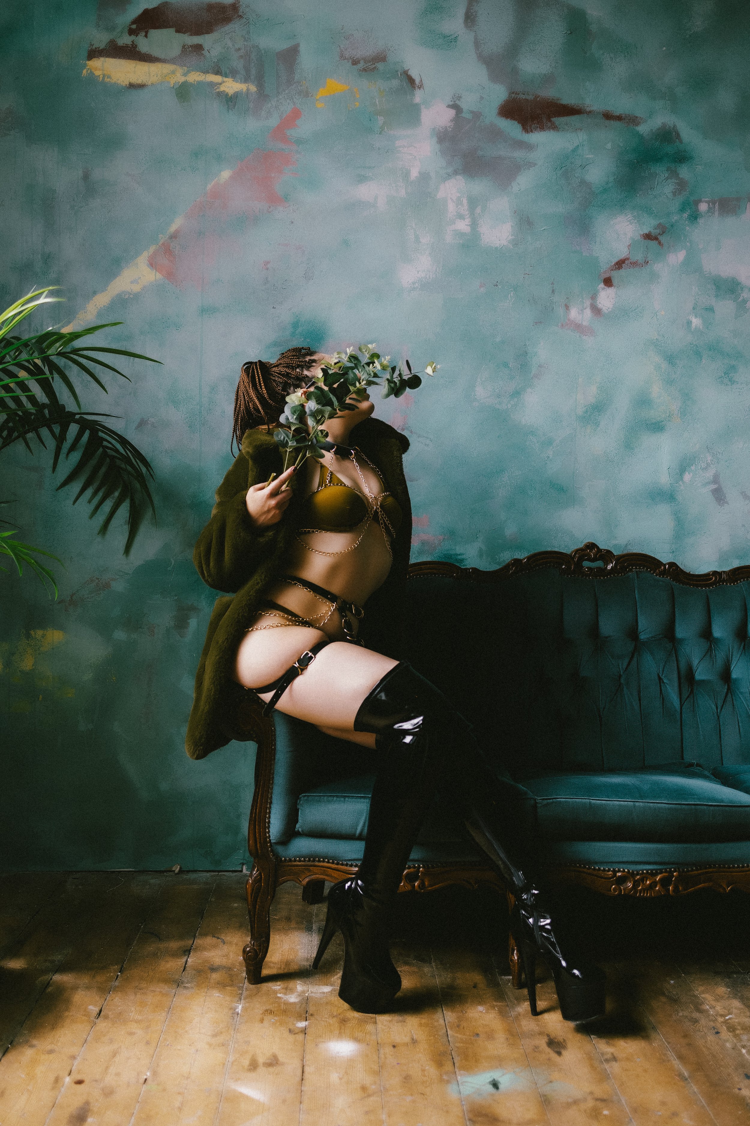 A woman in lingerie, thigh-high black boots, and a fur coat sitting on a vintage teal sofa, holding a leafy branch in front of her face, with a distressed teal wall and a potted plant in the background.