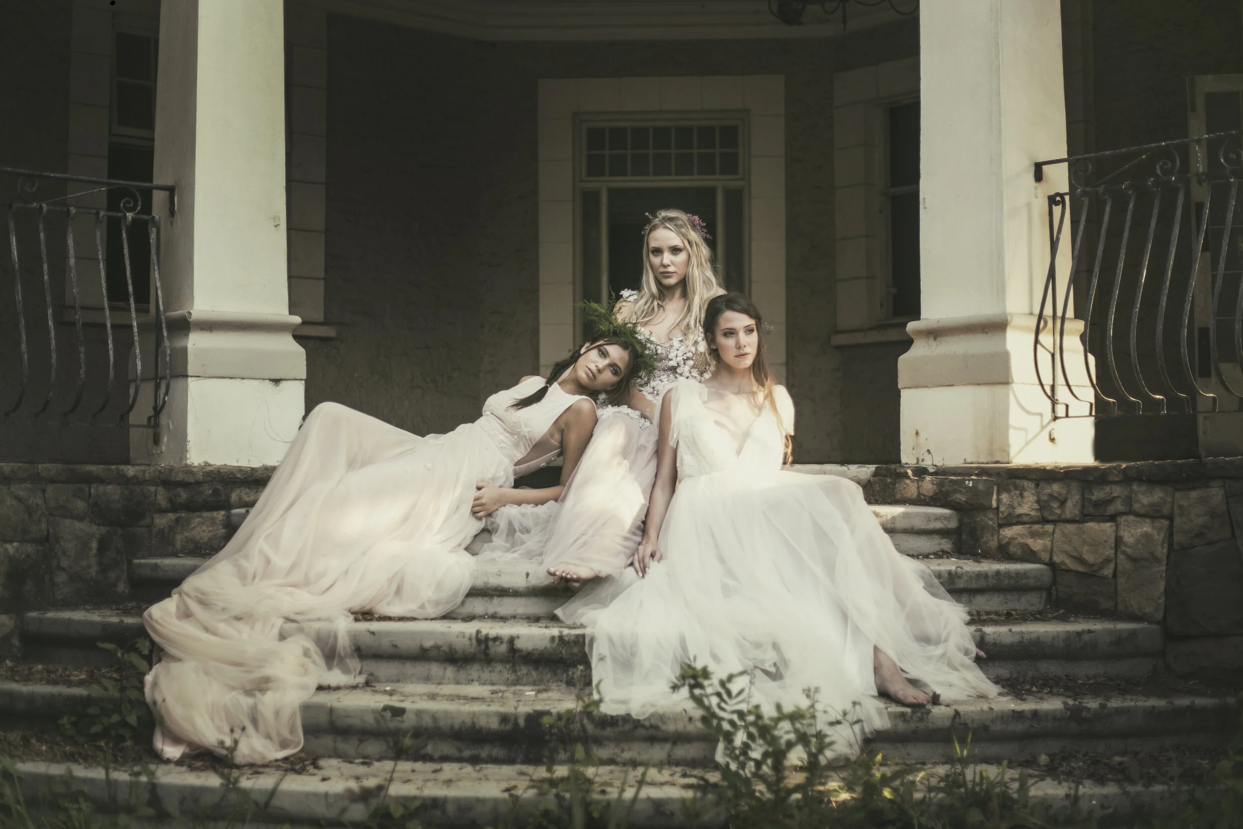 Four women in flowing white dresses pose on the steps of an old building with stone and stucco exterior, overgrown plants at the bottom.