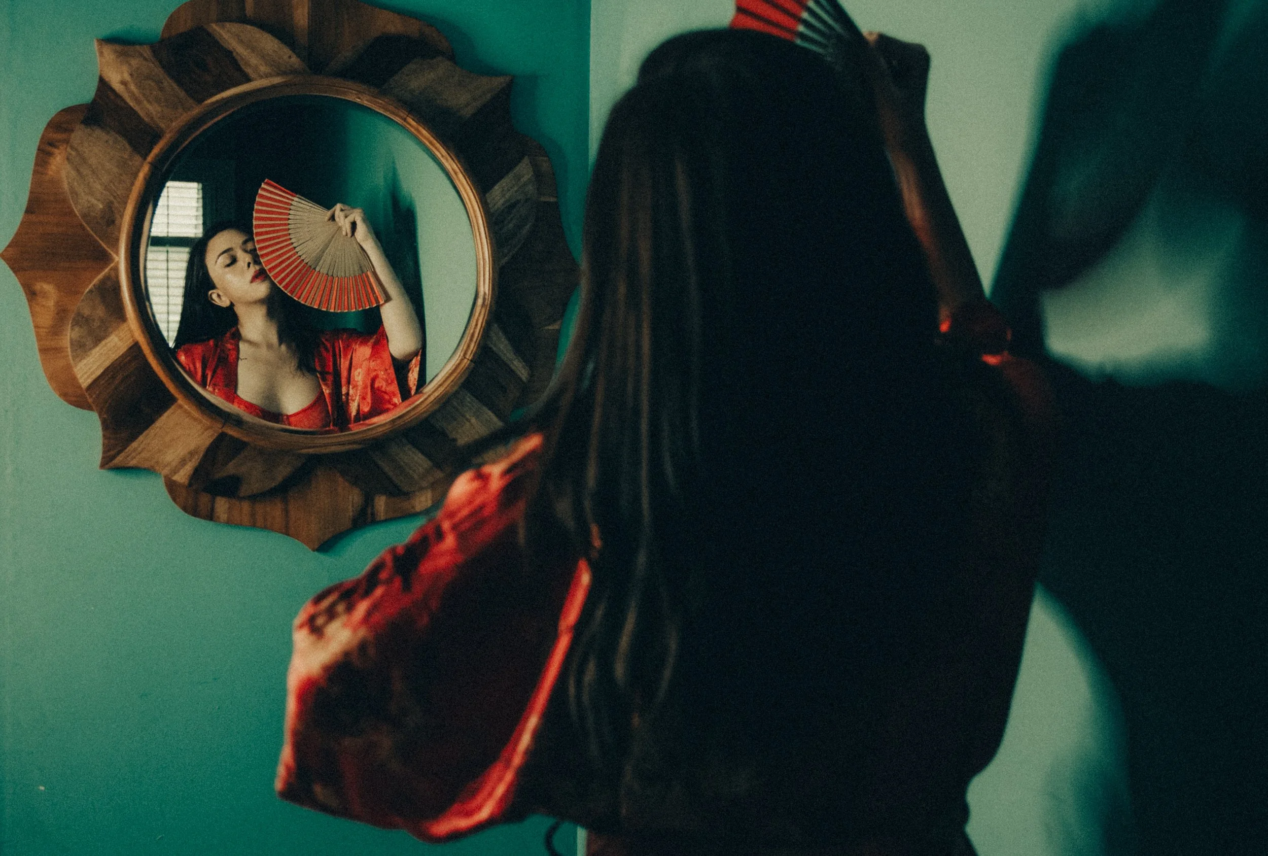 A woman in black clothes and a red and black patterned sleeve is looking at her reflection in a round mirror with a wooden frame. She is holding a red and gold folding fan, partially covering her face. The mirror is mounted on a teal wall.