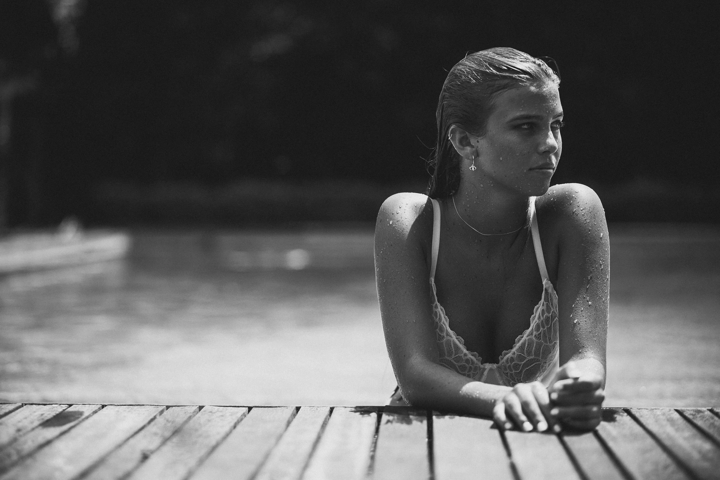 A woman in a swimsuit leaning on a dock by the water, with wet hair and water droplets on her skin, looking to the side in black and white.