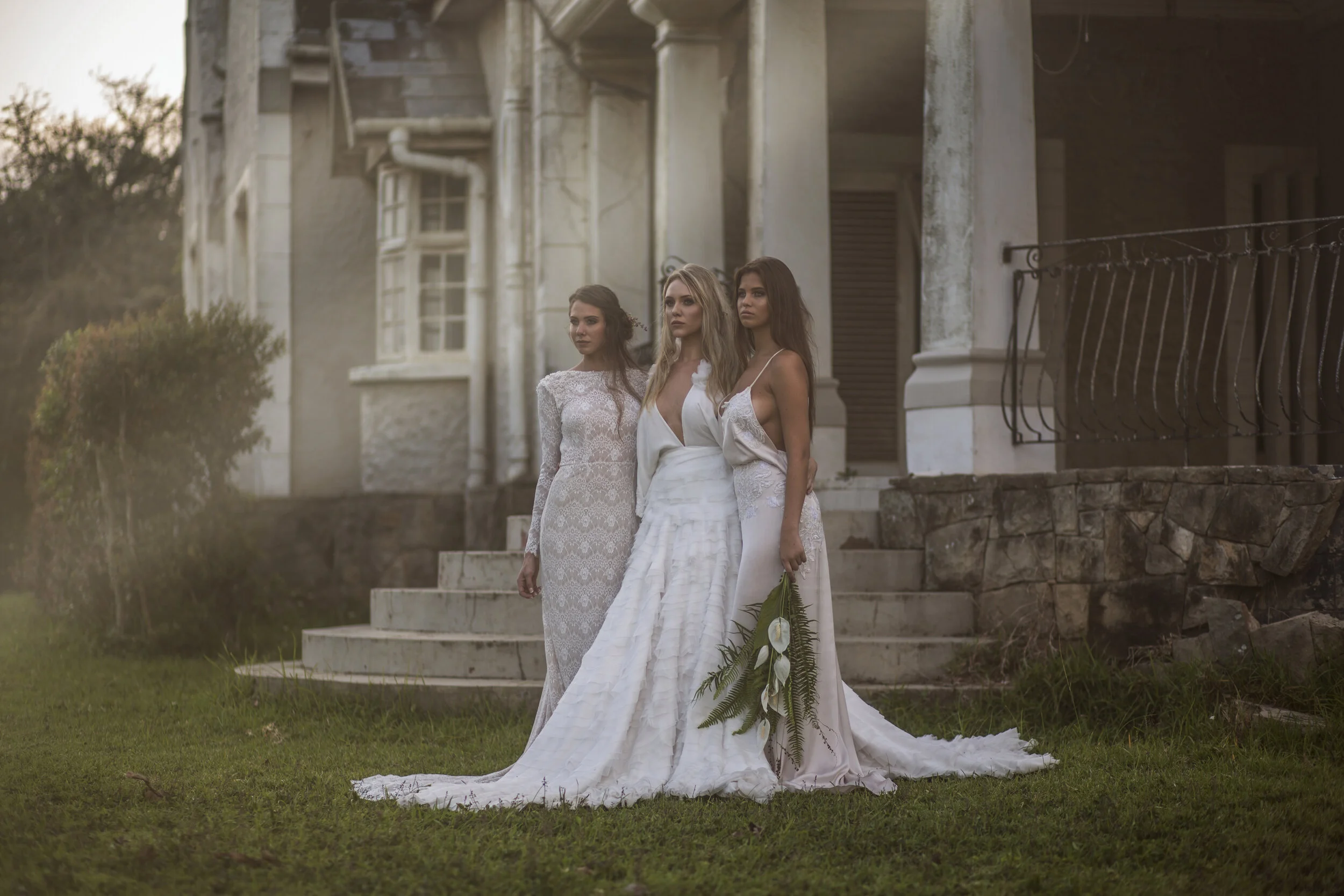 Three women in white dresses stand on grass in front of a large, old building during sunset. One woman holds a bouquet of white flowers and greenery.