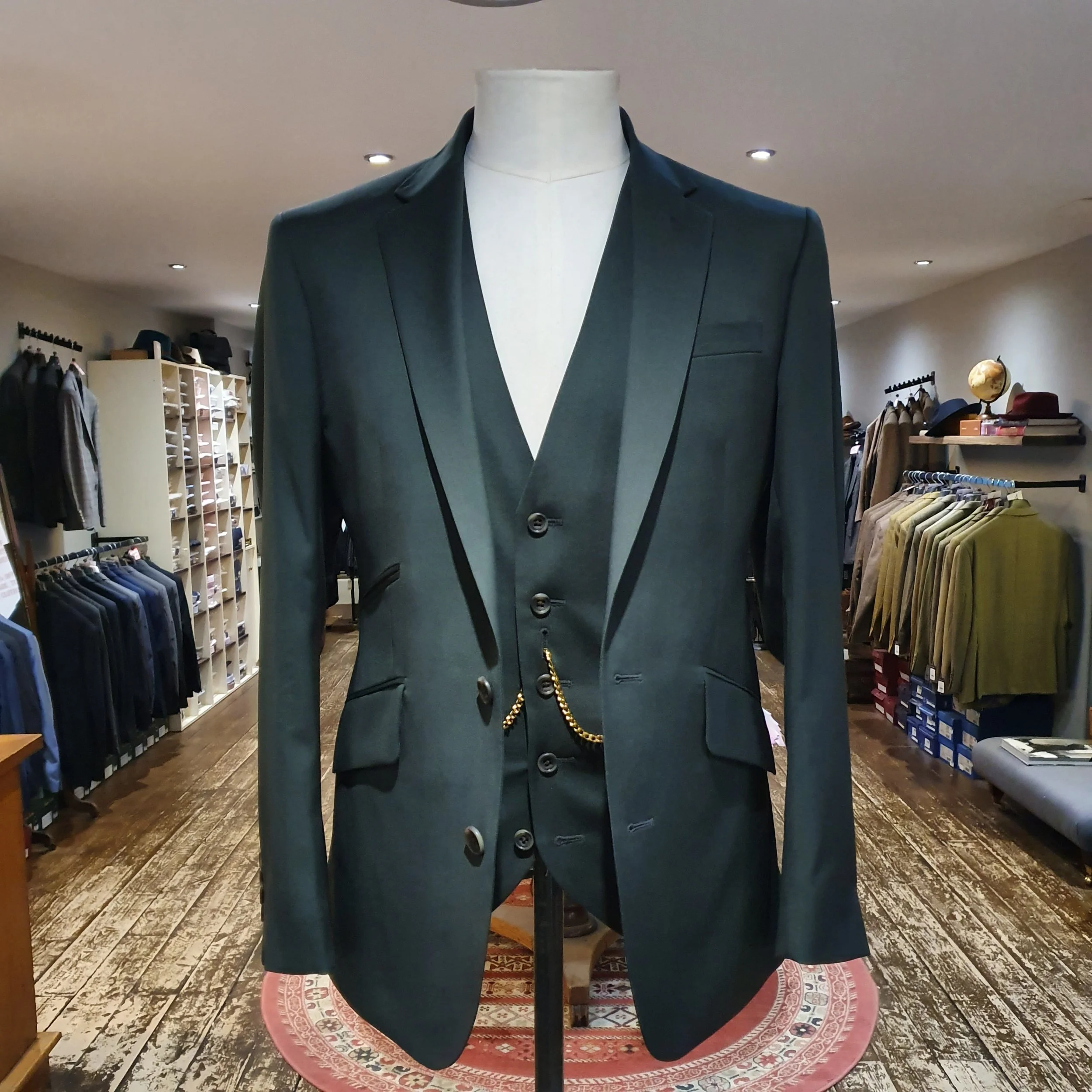 Mannequin dressed in a dark green suit with a vest and a chain, inside a clothing store with shelves of folded clothes and racks of jackets in the background.