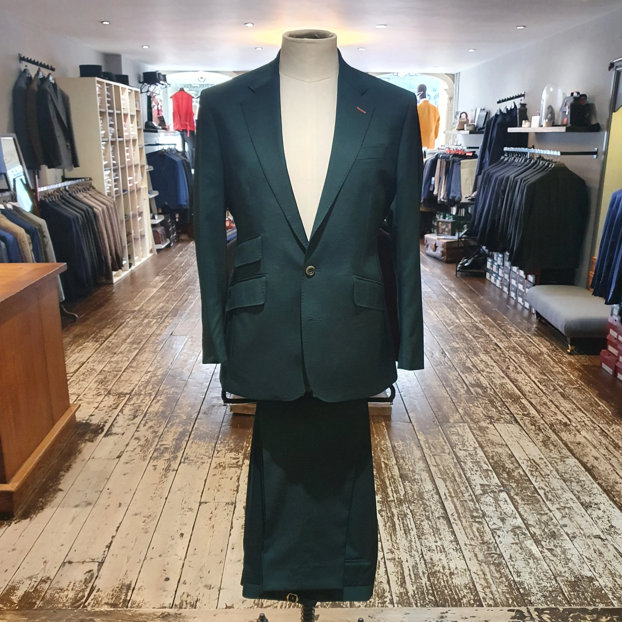 Mannequin wearing a dark green suit jacket over a light-colored turtleneck, in a clothing store with racks of suits and clothing in the background.