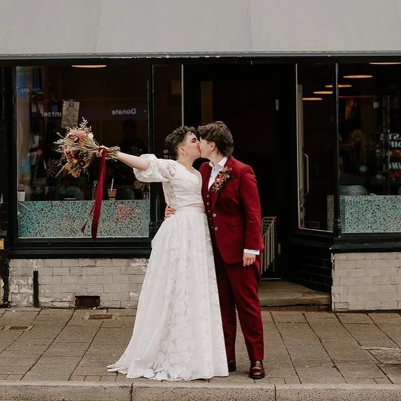 A bride and groom share a kiss outside a building, with the bride holding a bouquet and her arm extended.