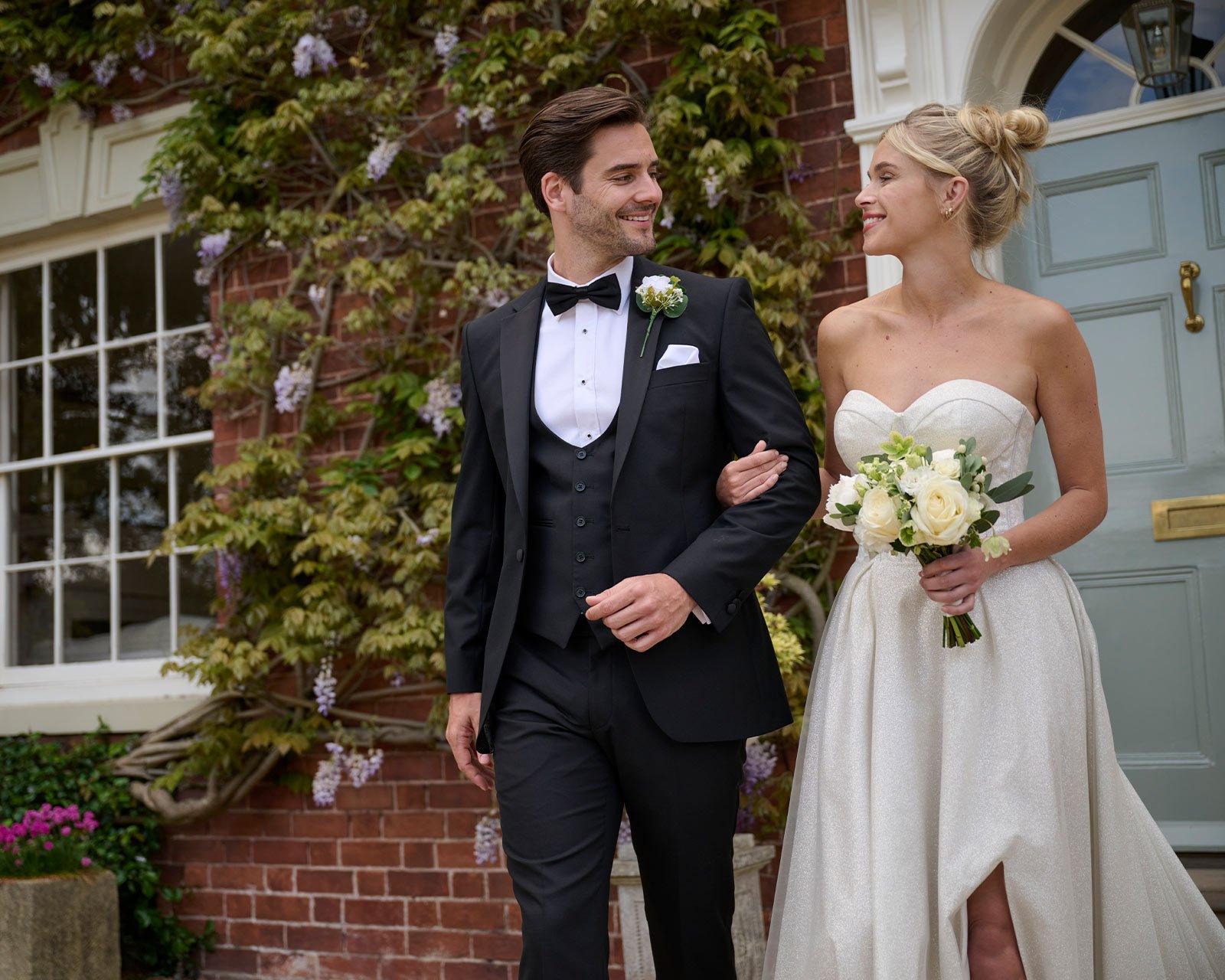 A bride and groom smiling and walking arm in arm outside a house decorated with climbing flowers. The groom is wearing a black tuxedo with a bow tie, and the bride is wearing a strapless white wedding gown holding a bouquet of white roses.
