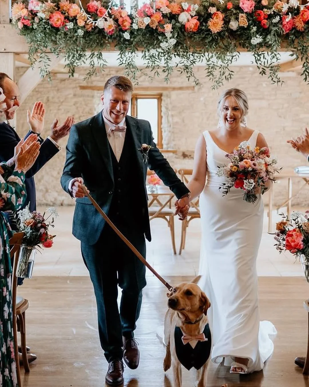 Wedding couple, a man in a tuxedo and a woman in a white wedding dress, holding hands and walking down the aisle, accompanied by a dog dressed in a tuxedo and bow tie, with guests clapping and smiling in a venue decorated with pink and white flowers and greenery.