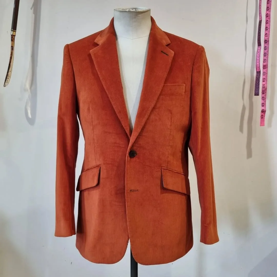 A single-breasted orange blazer with notch lapels, pockets, and a button, displayed on a dress form. In the background, there is a pink measuring tape hanging on the wall.