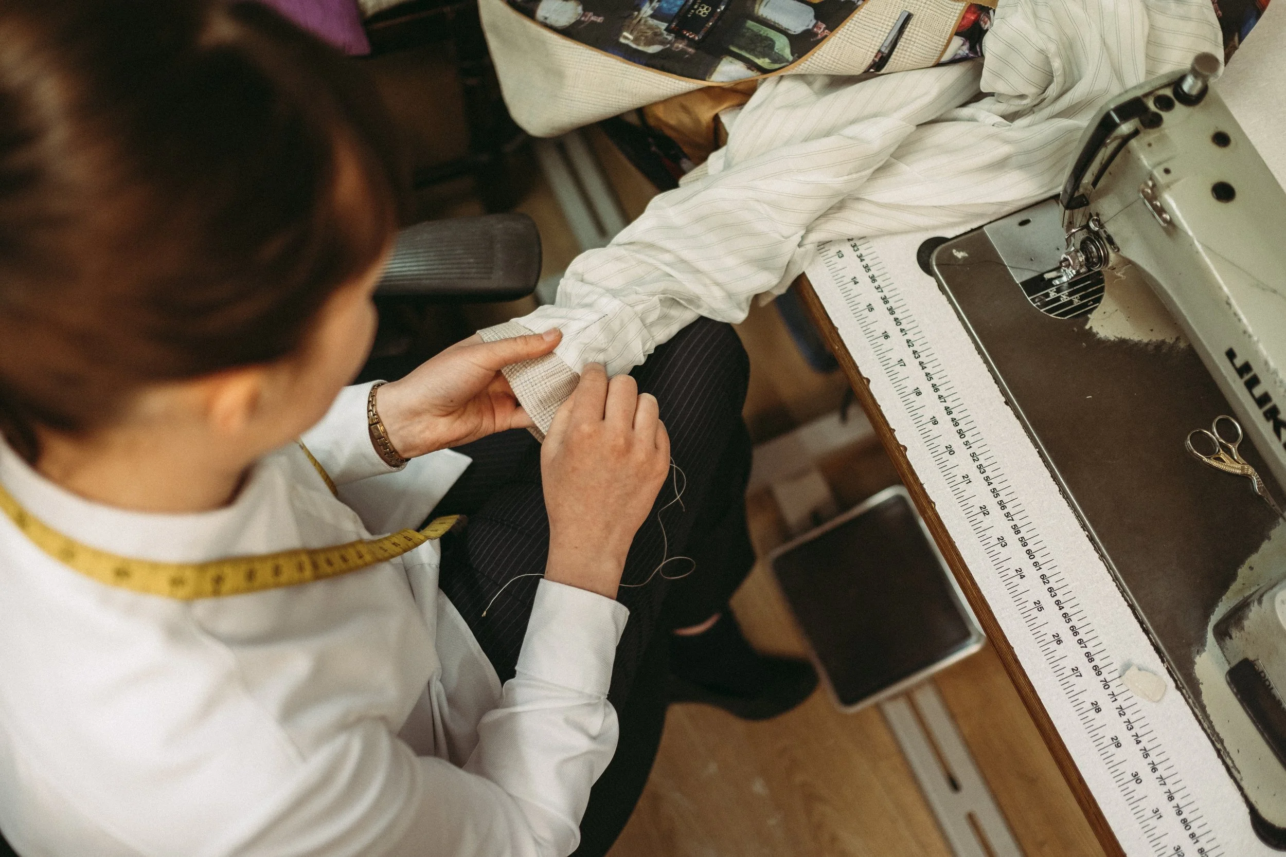 Seamstress sewing a cuff on a white shirt at a work table with measuring tape, sewing machine, and scissors.