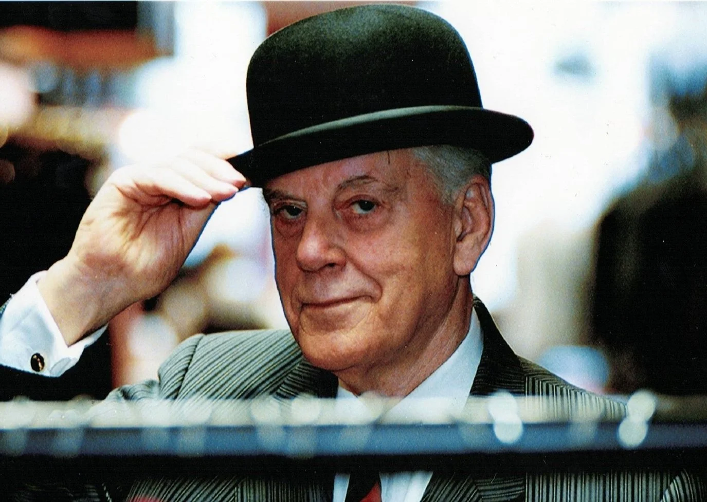 Elderly man wearing a bowler hat, tilting it with his hand, dressed in a suit.