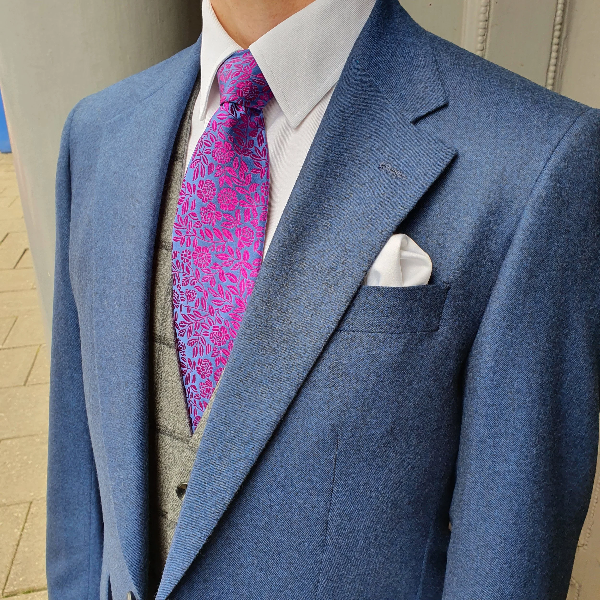 Close-up of a man's chest wearing a blue suit jacket, white dress shirt, pink patterned tie, gray vest, and white pocket square.