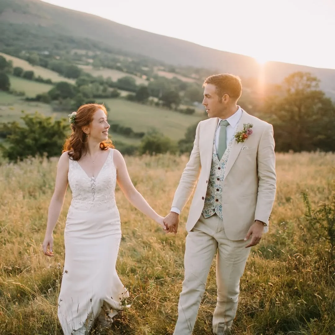 A bride and groom holding hands in a grassy field during sunset, with rolling hills in the background.