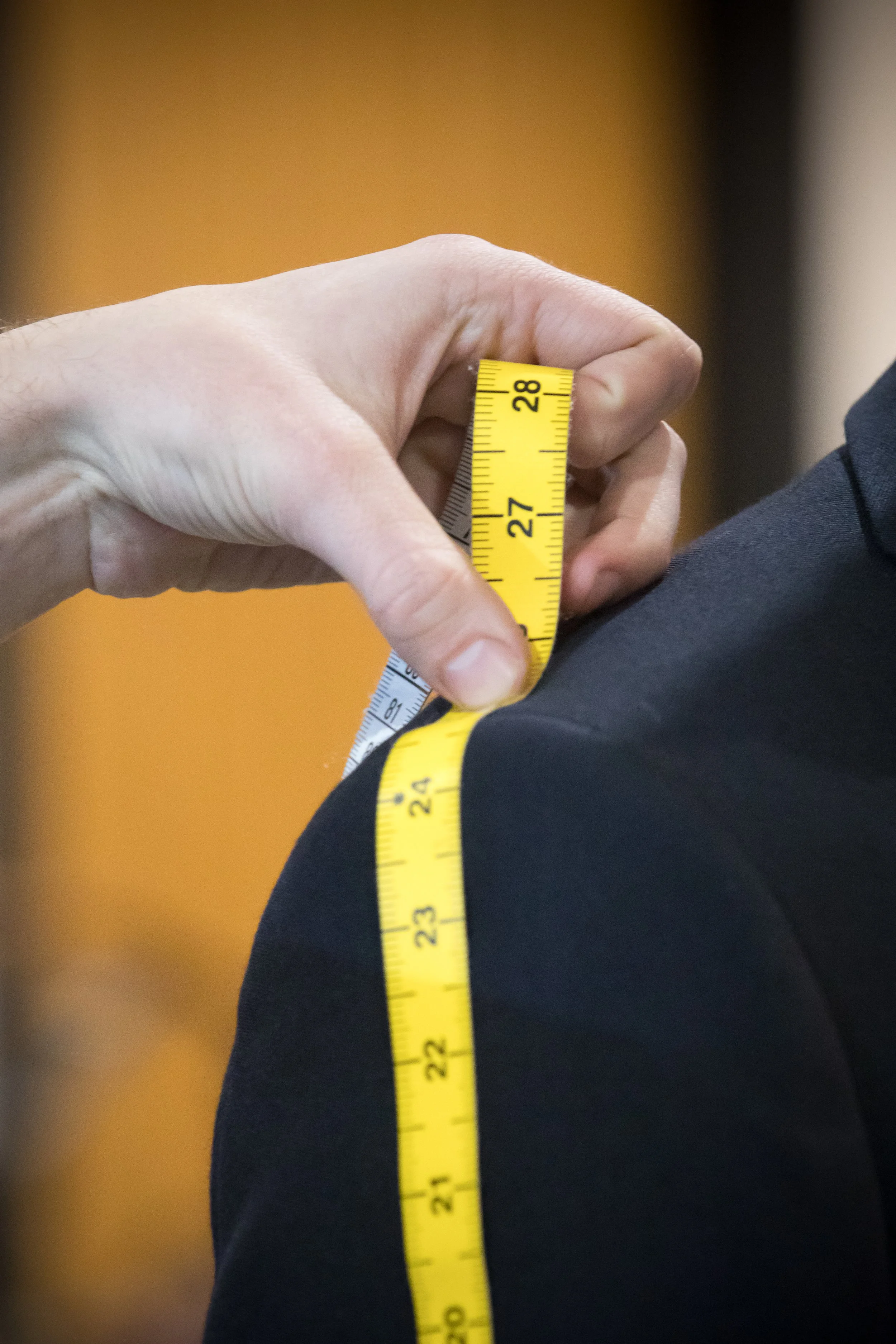 A person using a yellow measuring tape to measure the shoulder seam of a dark-colored suit jacket.