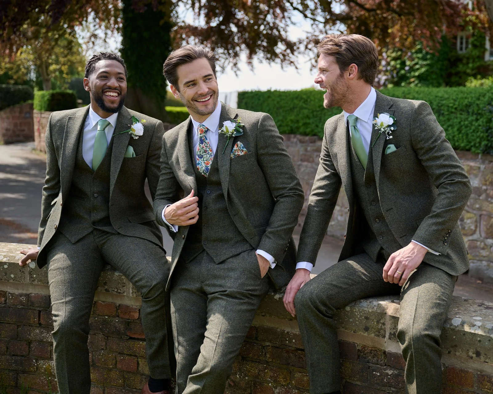 Three men in suits sitting and standing on a brick wall, laughing and talking outdoors on a sunny day with trees and greenery in the background.
