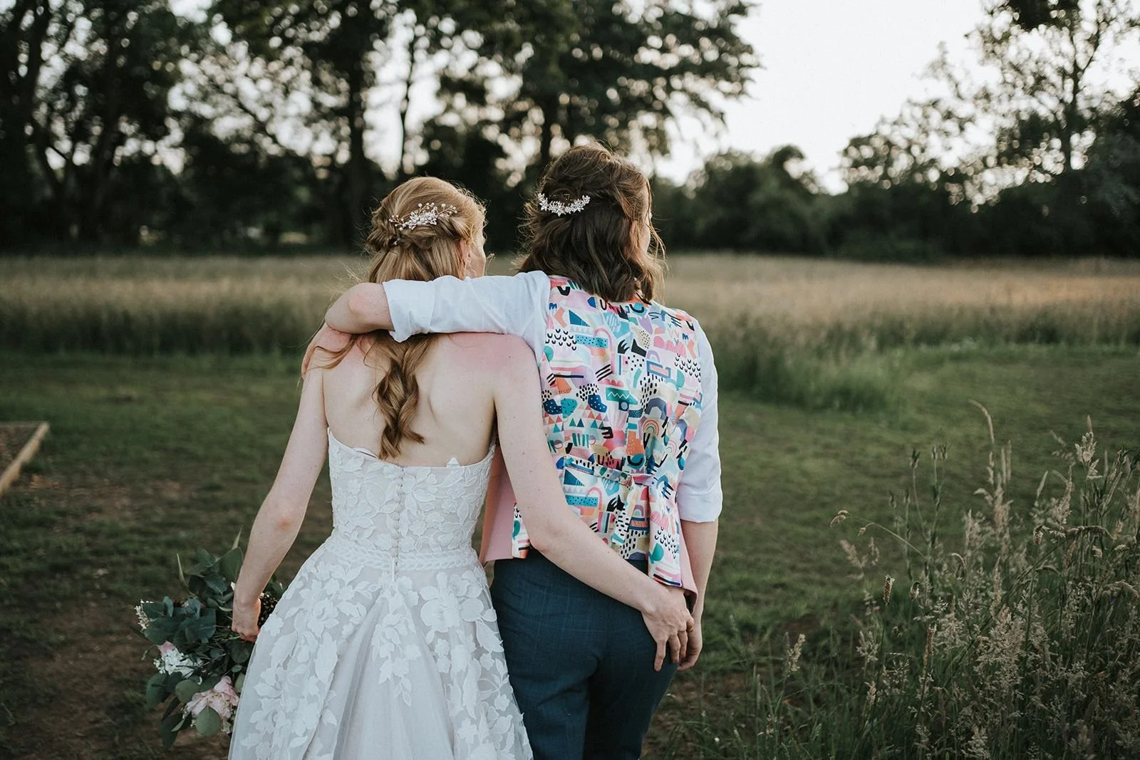Two women, one dressed in a white wedding gown holding a bouquet, and the other in a colorful patterned blazer and dark pants, are standing outdoors at sunset, with their arms around each other, overlooking a field with trees in the background.