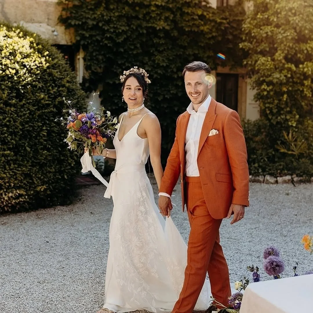 A couple dressed in wedding attire walking outdoors, smiling, with a woman holding a bouquet of flowers and a man in an orange suit. The background shows greenery and warm sunlight.